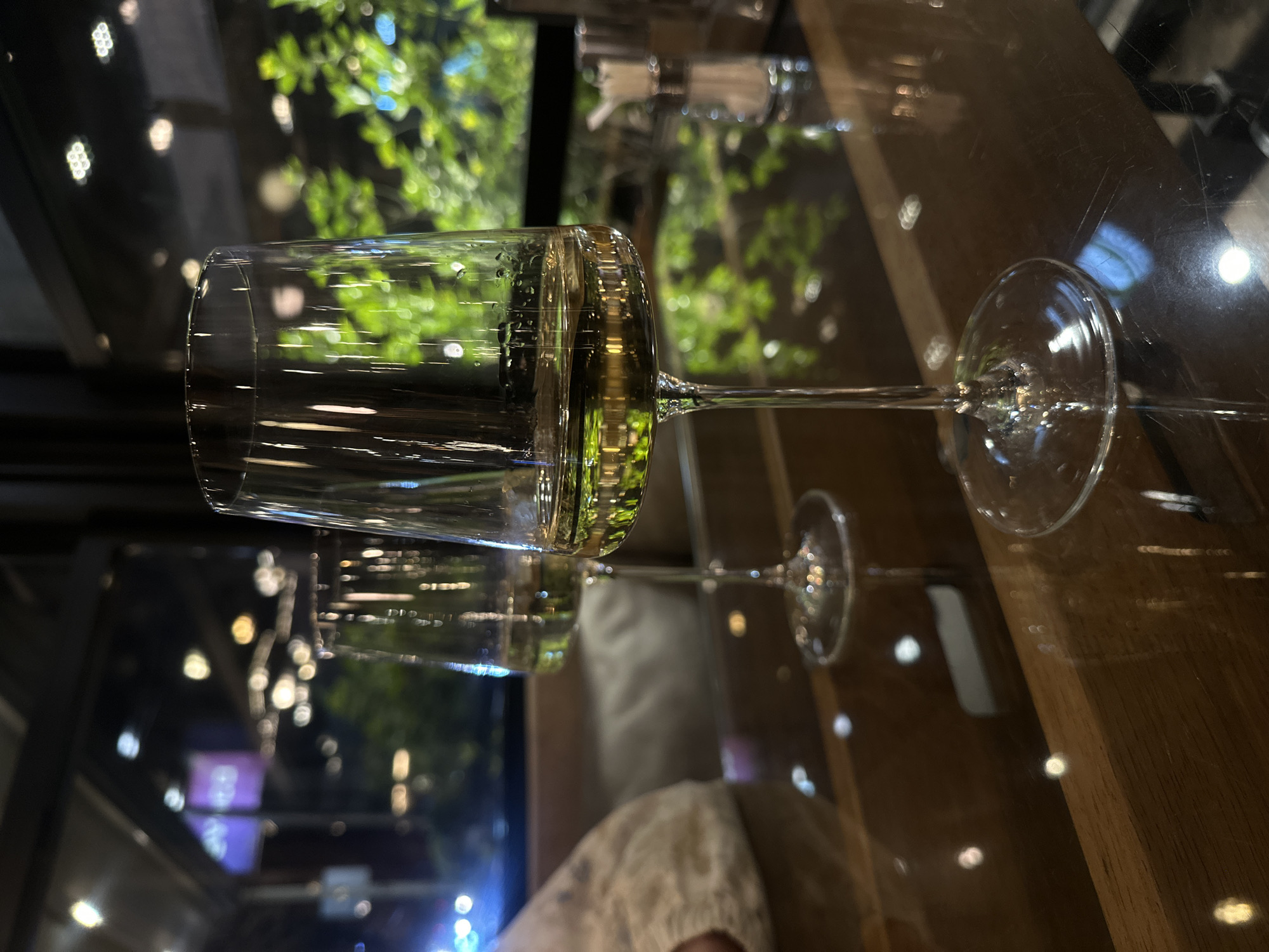 A glass of wine is placed horizontally on a table. A reflection of the glass and its contents is visible on the surface of the table. The background showcases an out-of-focus urban landscape with greenery and bokeh lights, potentially reflecting Yerevan, Armenia at night. The environment suggests a restaurant or bar setting. No people are visible in the immediate frame, though a hint of someone's clothing can be seen.