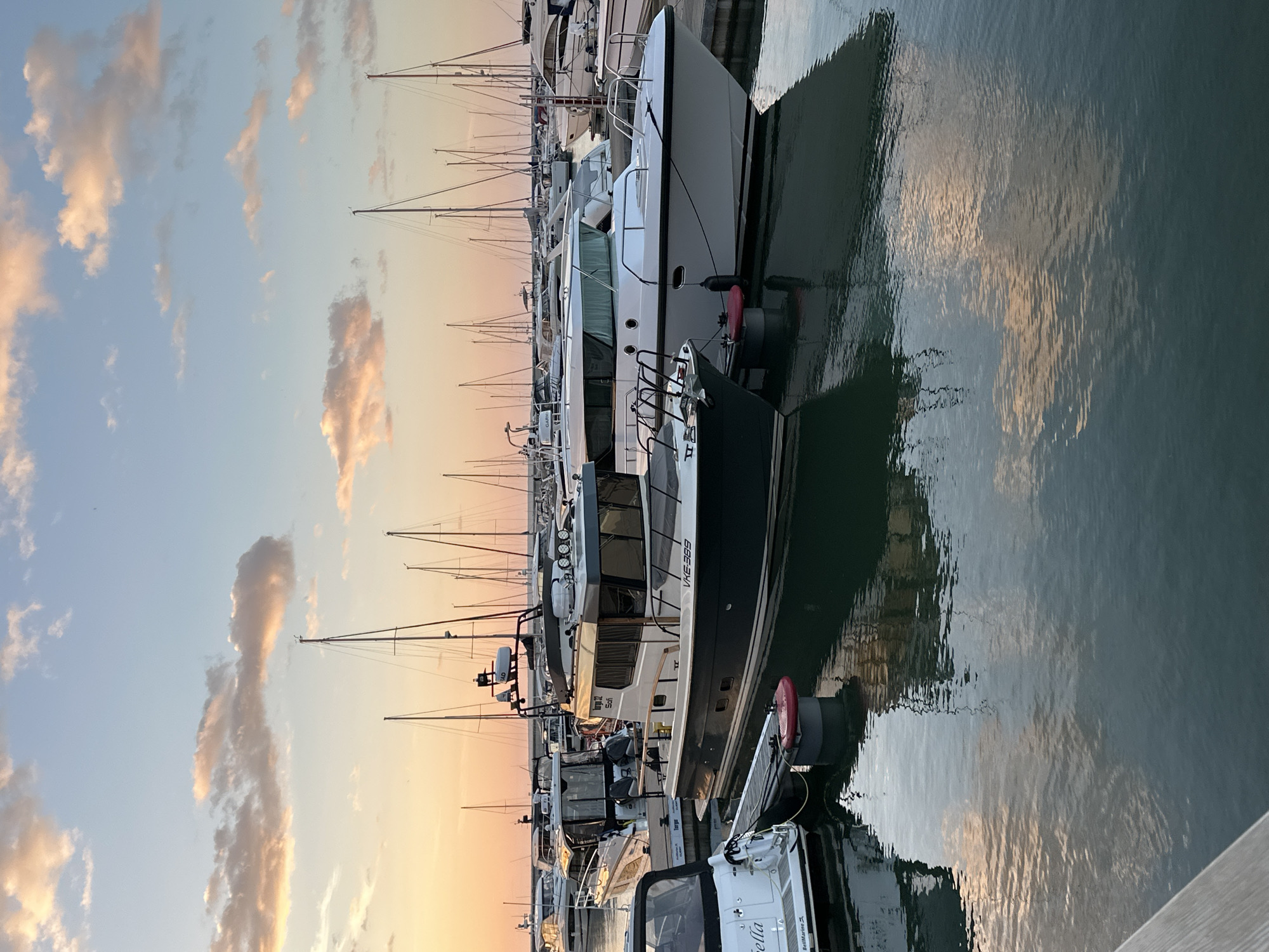 This is a photograph of a marina in Tallinn, Estonia, at sunset. The sky is a soft gradient of blue and orange, with scattered clouds that reflect the warm light of the setting sun. Several boats are docked in the water, their reflections shimmering on the surface. In the foreground, there is a dark blue and white motorboat with a cabin. To its right, a larger white motorboat is docked. In the background, numerous sailboats with tall masts are visible, as are other motorboats. The water is calm, with gentle ripples. A wooden pier is visible in the lower left corner of the image. The scene conveys a sense of tranquility and the beauty of a coastal evening.