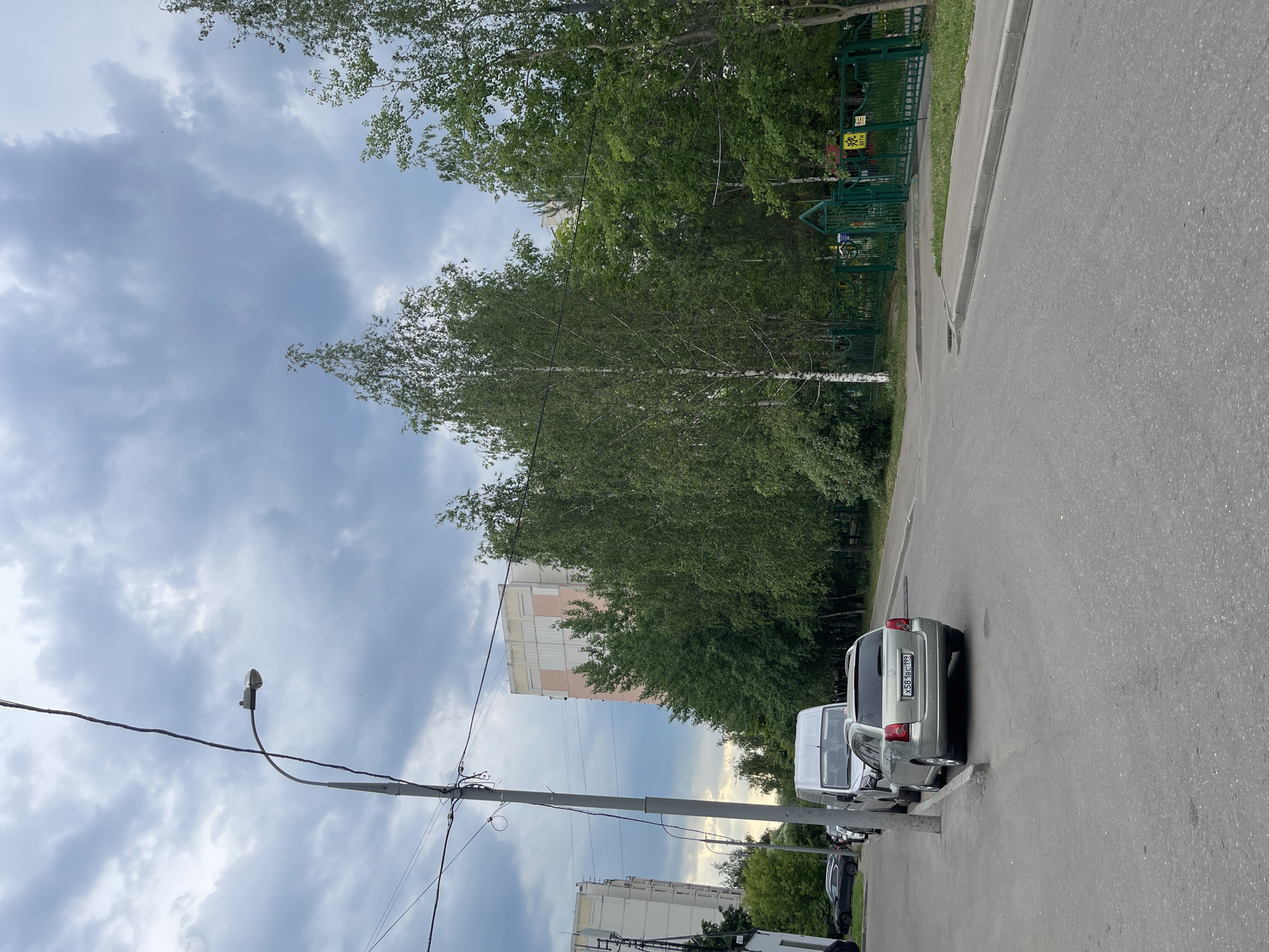 The image depicts a street scene in Moscow, Russia, under a cloudy sky. In the foreground, a gray asphalt road curves to the right, bordered by a concrete curb. Parked along the left side of the road are several vehicles, including a silver station wagon with a license plate displaying "X581BC 199" and a white van. Behind the parked cars, a tall utility pole with electrical wires stretches across the frame. In the middle ground, a row of trees, including birch trees with distinctive white bark, lines the street. Beyond the trees, a green metal fence encloses an area with a yellow sign featuring children and the word "ДЕТИ" (children). In the background, two multi-story apartment buildings with light-colored facades are visible. The overall environment suggests a residential urban area.