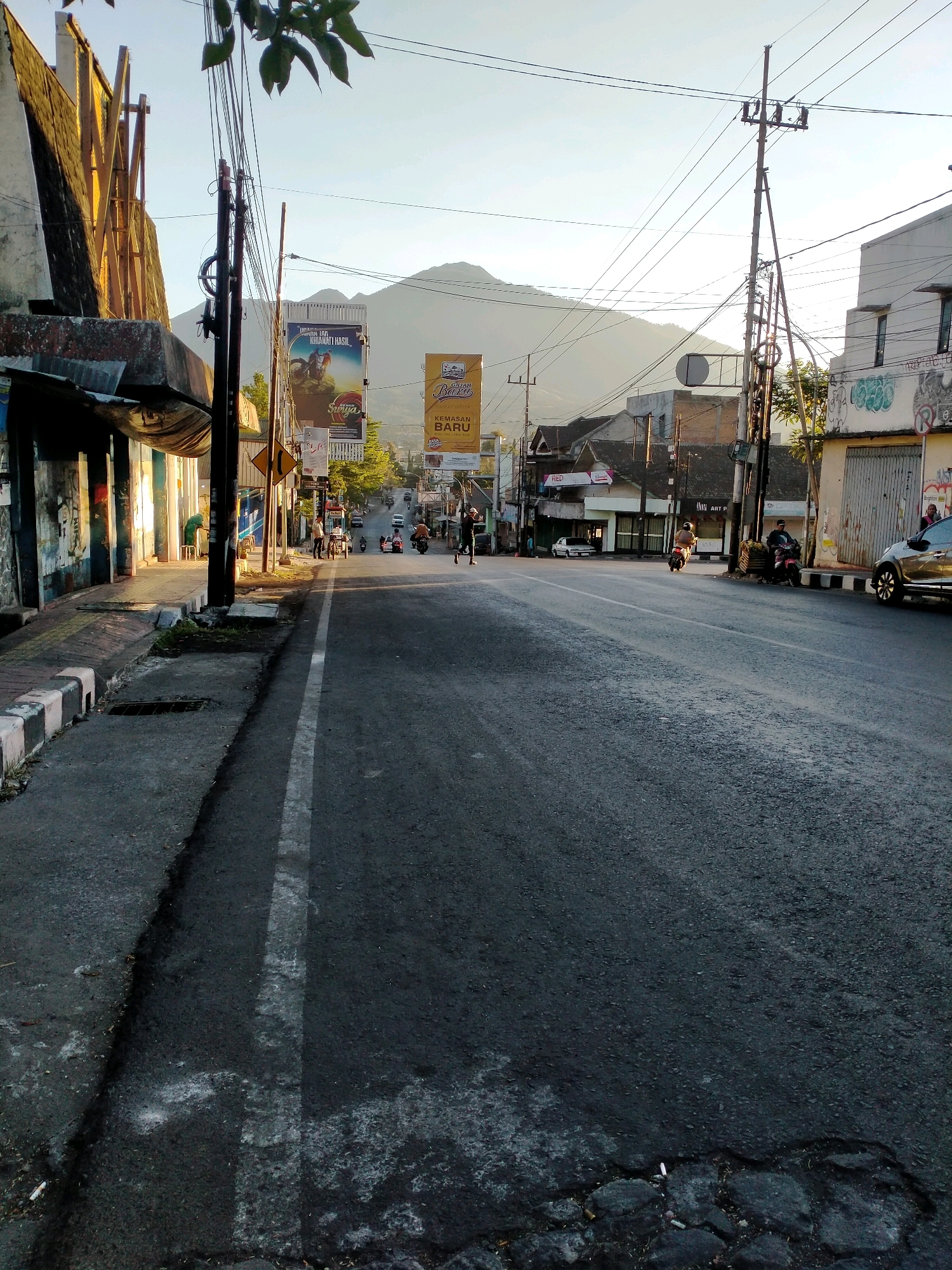 The image captures a street scene in Batu, Indonesia, likely during early morning or late afternoon, given the soft lighting and long shadows. The perspective is from the side of the road, looking forward down a paved street.
On the left side of the street, there is a sidewalk with a covered area supported by columns. The structure above appears weathered and possibly abandoned, with a corrugated metal roof and exposed beams. Graffiti is visible on some of the columns. The sidewalk is bordered by a curb with alternating black and white painted sections, and a grate is set into the pavement.
The street itself is asphalt, with a white line painted down the center. There is evidence of wear and tear on the road surface, with some patches and cracks visible.
Along the right side of the street, there are various buildings. One building has a large, closed metal gate and graffiti on its wall. Further down, more buildings are visible, some with multiple stories. Several motorcycles are parked or being ridden on the street. Two individuals are visible on a motorcycle carrying a load of what appears to be produce. Another person is standing near a motorcycle parked on the side of the road. A silver car is parked on the right side of the street.
Dominating the background is a large, green mountain range, partially obscured by the buildings and trees. The sky is clear and bright, indicating favorable weather conditions. Several billboards are placed along the street, featuring advertisements in Indonesian.
The scene depicts a typical urban street in Indonesia, with a blend of residential or commercial buildings, infrastructure elements like power lines and poles, and everyday activities like commuting and local commerce. The presence of the mountain range suggests the location is in or near a mountainous region, consistent with Batu's geography.