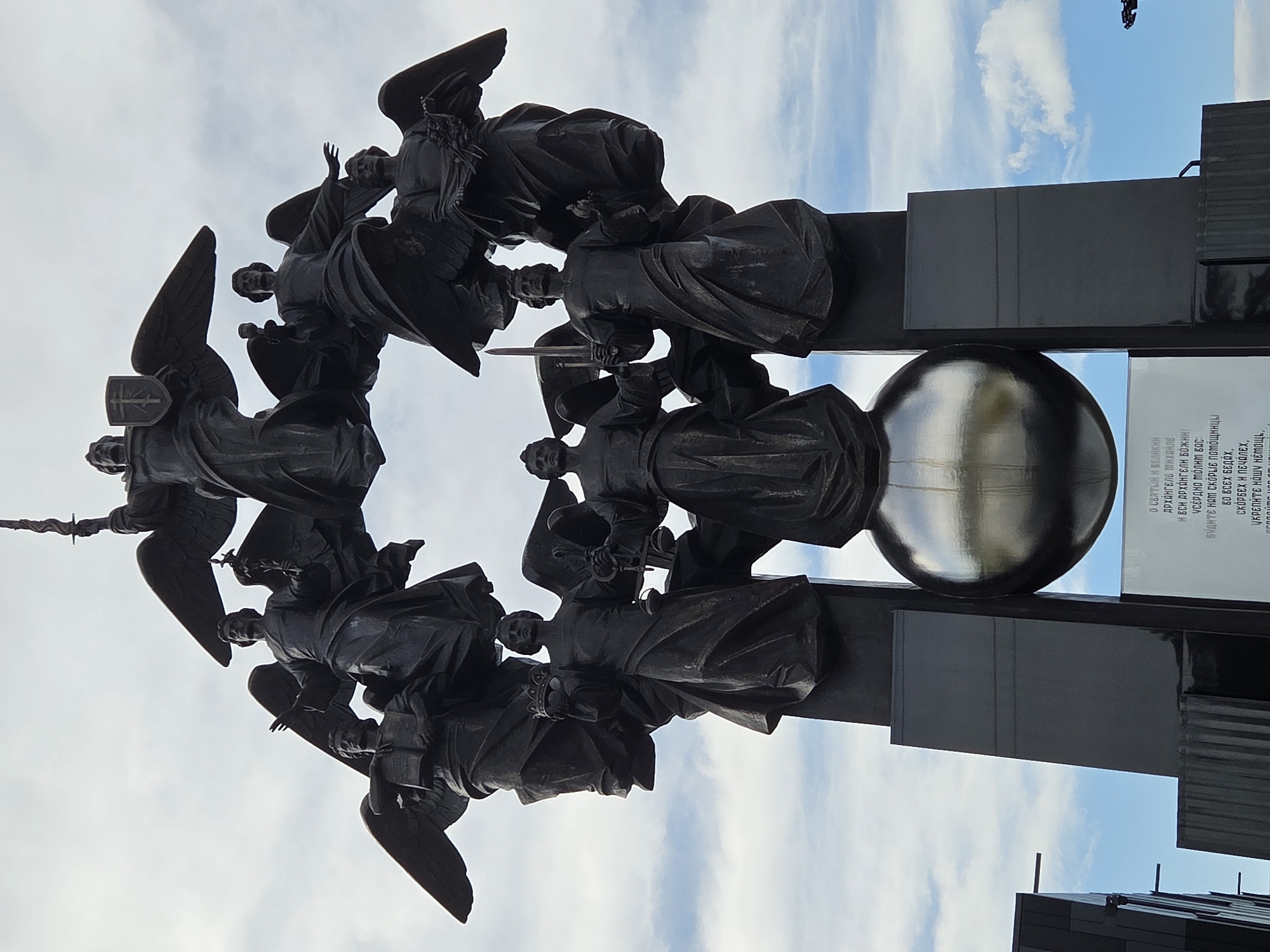 The image shows the monument "The Archangel Michael with the Archangels" located in Yekaterinburg, Russia. The monument is a bronze sculpture of an archangel at the top, surrounded by other winged figures. The archangel at the top is holding a sword and a shield with a cross. The other figures are holding various objects, including a book, a crown, a laurel wreath, and a scale. The sculpture is mounted on a pedestal that includes a large silver sphere. Below the sphere is a white marble plaque with text inscribed on it. The monument is set against a backdrop of a cloudy sky.