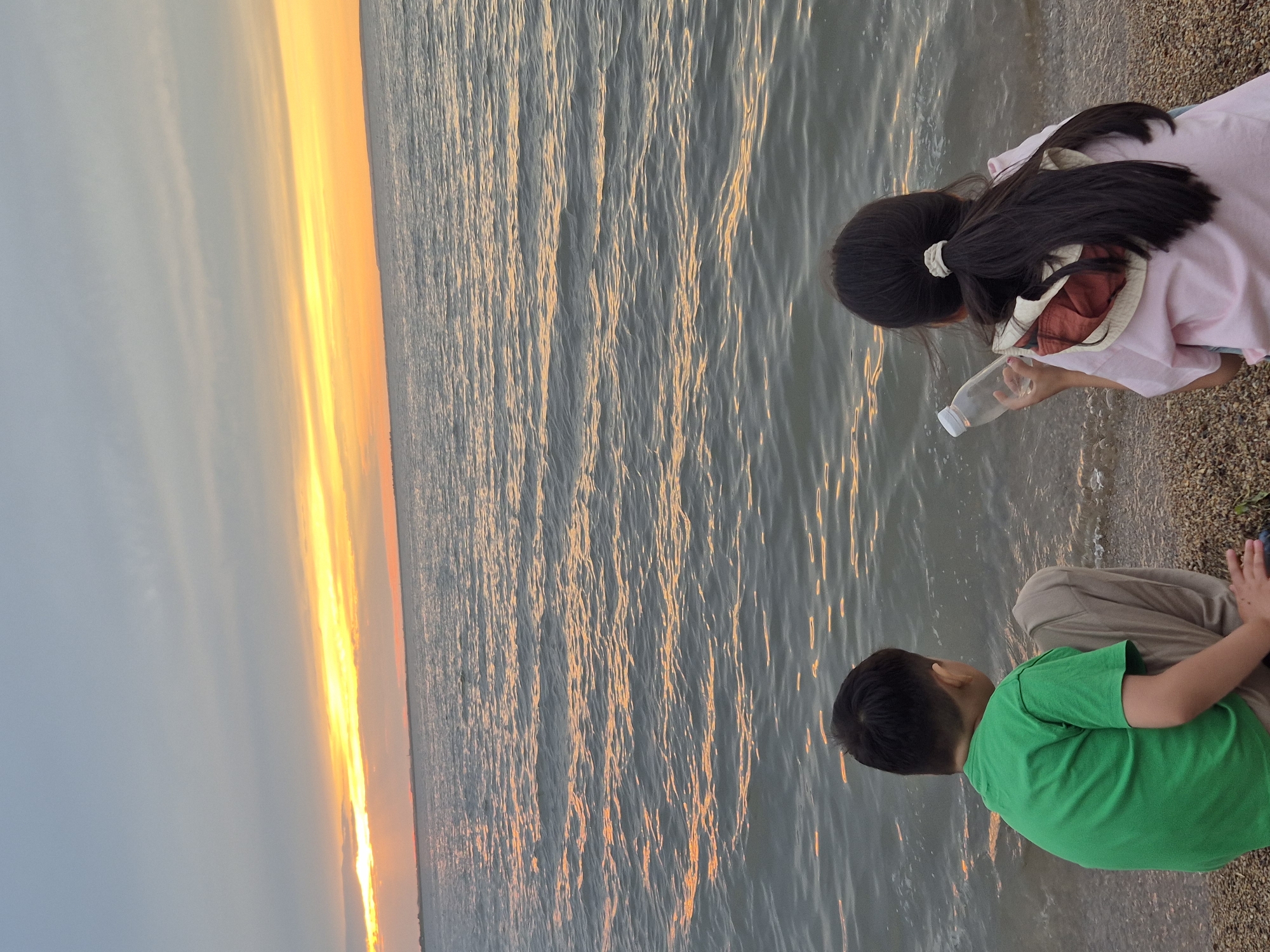 The image depicts two children by a body of water at sunset. A girl with long dark hair tied back with a white scrunchie is wearing a pink shirt and is crouched by the water's edge, holding a clear plastic bottle. Next to her, a boy wearing a bright green t-shirt and khaki pants is also crouched down, facing away from the camera. The water has a rippling surface reflecting the warm orange and yellow hues of the setting sun. The sky transitions from a soft blue to vibrant yellow and orange near the horizon, indicating it is either sunrise or sunset. The ground where the children are sitting appears to be a mix of sand and small pebbles. The overall scene conveys a peaceful moment of children interacting with nature by the water. The location is identified as Kokshetau, Kazakhstan. There is no visible text in the image.