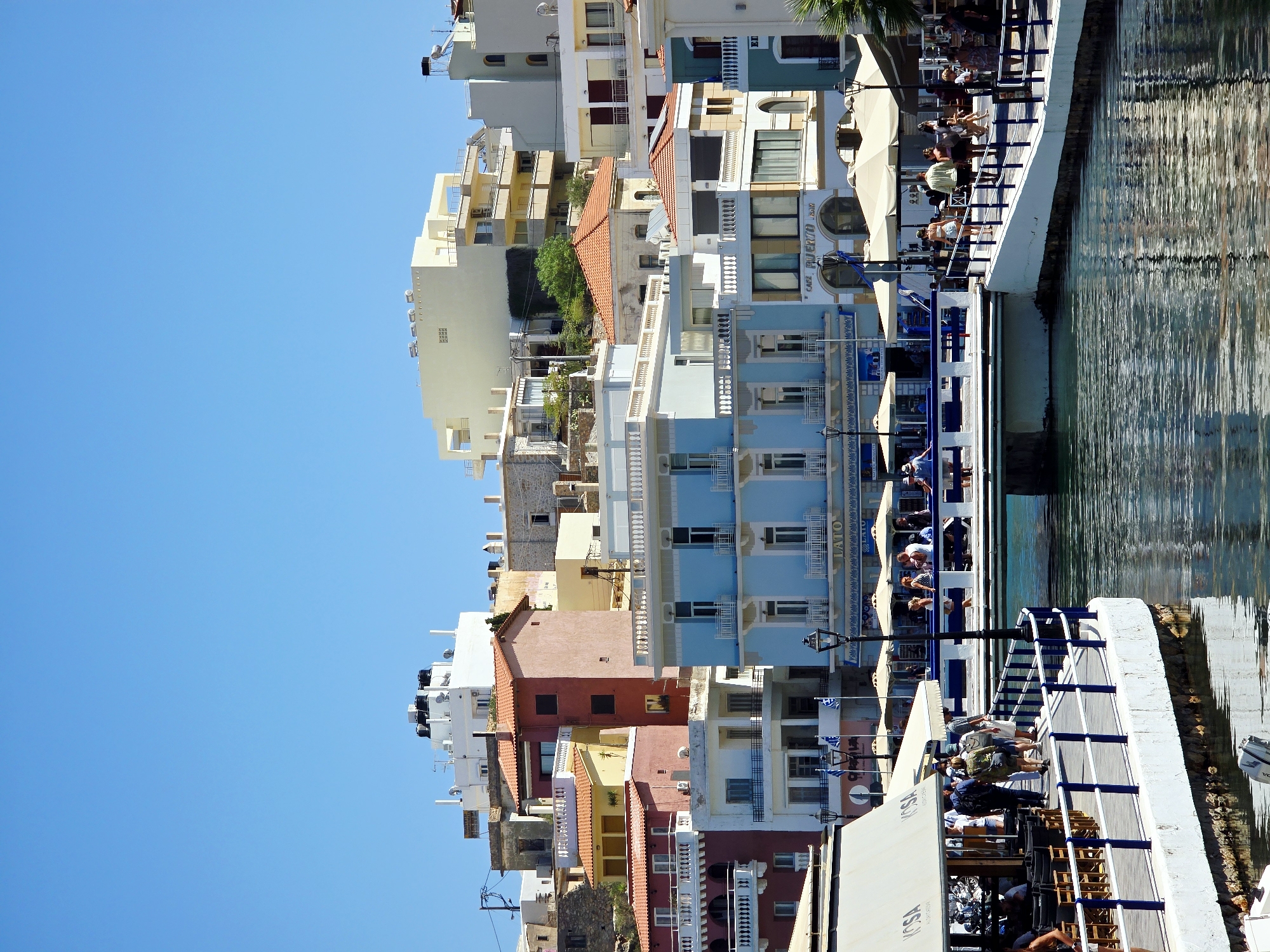 The image captures a bustling waterfront scene in Agios Nikolaos, Greece, under a clear blue sky. The foreground features a broad, white promenade with a dark blue railing, where several people are walking and socializing. Umbrellas are set up along the promenade, suggesting outdoor dining or seating areas. On the left, a building with a white awning, labeled "KOSA," appears to be a restaurant or cafe, with tables and chairs visible underneath.
Across the water, a picturesque view of Agios Nikolaos unfolds. Several multi-story buildings in varying architectural styles and colors line the waterfront. Prominent among them is a striking light blue building with balconies and large windows. To its right, a cream-colored building displays arched entrances with signage that reads "CAFE PUERTO BAR". Further back, more buildings rise up the hillside, showcasing terracotta roofs and pastel facades, interspersed with more modern structures. The water itself is a deep blue, reflecting the surrounding buildings and the sky, with ripples indicating a gentle movement. The overall atmosphere is lively and inviting, characteristic of a popular Mediterranean tourist destination.