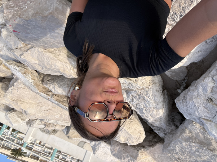 An adult female is captured in a close-up selfie-style shot on a rocky shoreline in Cannes, France. She is positioned mid-face to chest, facing right with a slight upward gaze and pursed lips. She has dark hair and wears a black, ribbed, short-sleeved crop top. Essential accessories include large, tortoise-shell patterned Cazal sunglasses with amber-tinted lenses and large, gold-colored hoop earrings. The immediate background consists of numerous large, light-colored, irregularly shaped rocks, characteristic of riprap. In the upper right background, a multi-story, light-colored building with balconies and a partial palm tree is visible. The scene is brightly illuminated by natural light, casting reflections on the sunglasses.