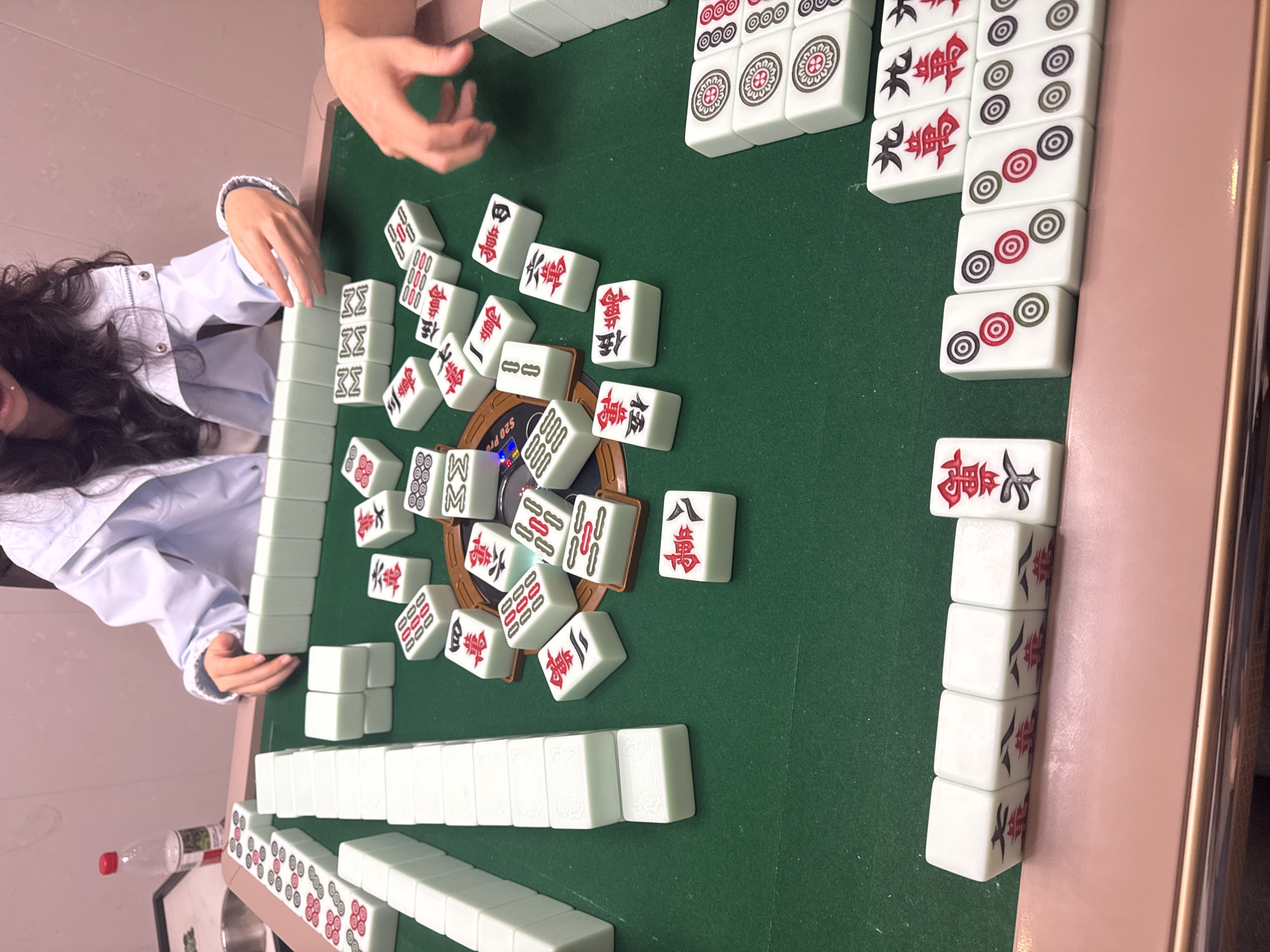 The image shows a mahjong game in progress. A person is shown on the left, wearing a white top. The game is set up on a green surface, presumably a mahjong table. Numerous white mahjong tiles are scattered and arranged on the table, displaying various Chinese characters and patterns in red and black ink. A round, brown object with "S20 Pro" printed is visible among the tiles, possibly a holder or a device. Additionally, stacks of organized tiles are positioned around the perimeter of the table.