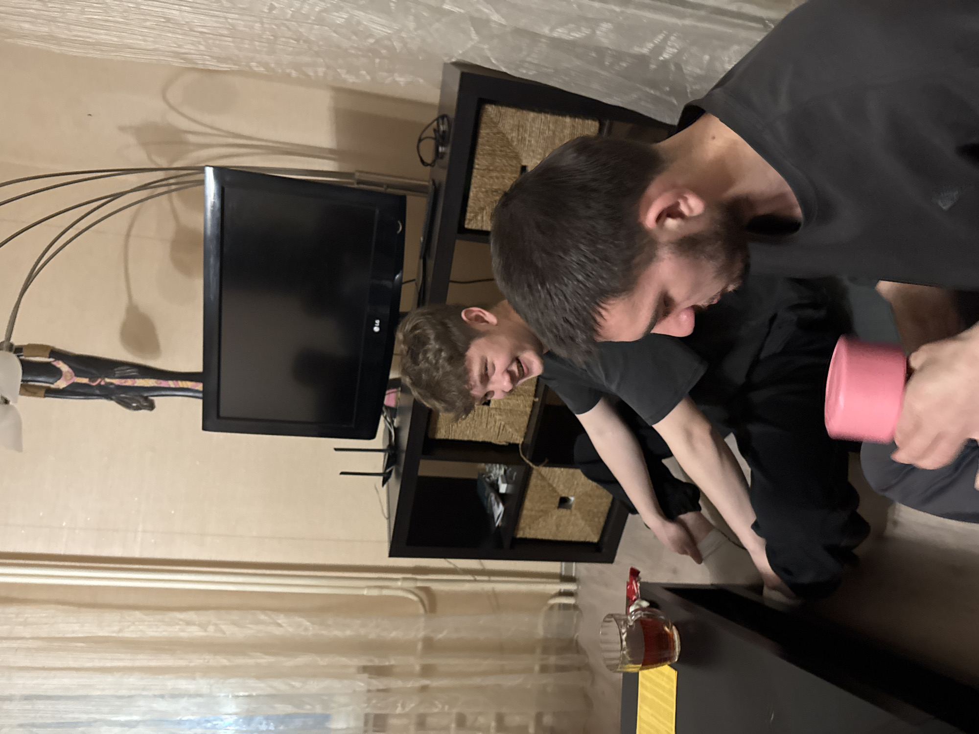 Two men are seated on the floor in an interior setting, likely a living room. A television is visible in the background, placed on a black entertainment center with wicker baskets. A cup and other items are on a black coffee table. The environment suggests a residential space. There is no location of Kazan, Russia in the image.