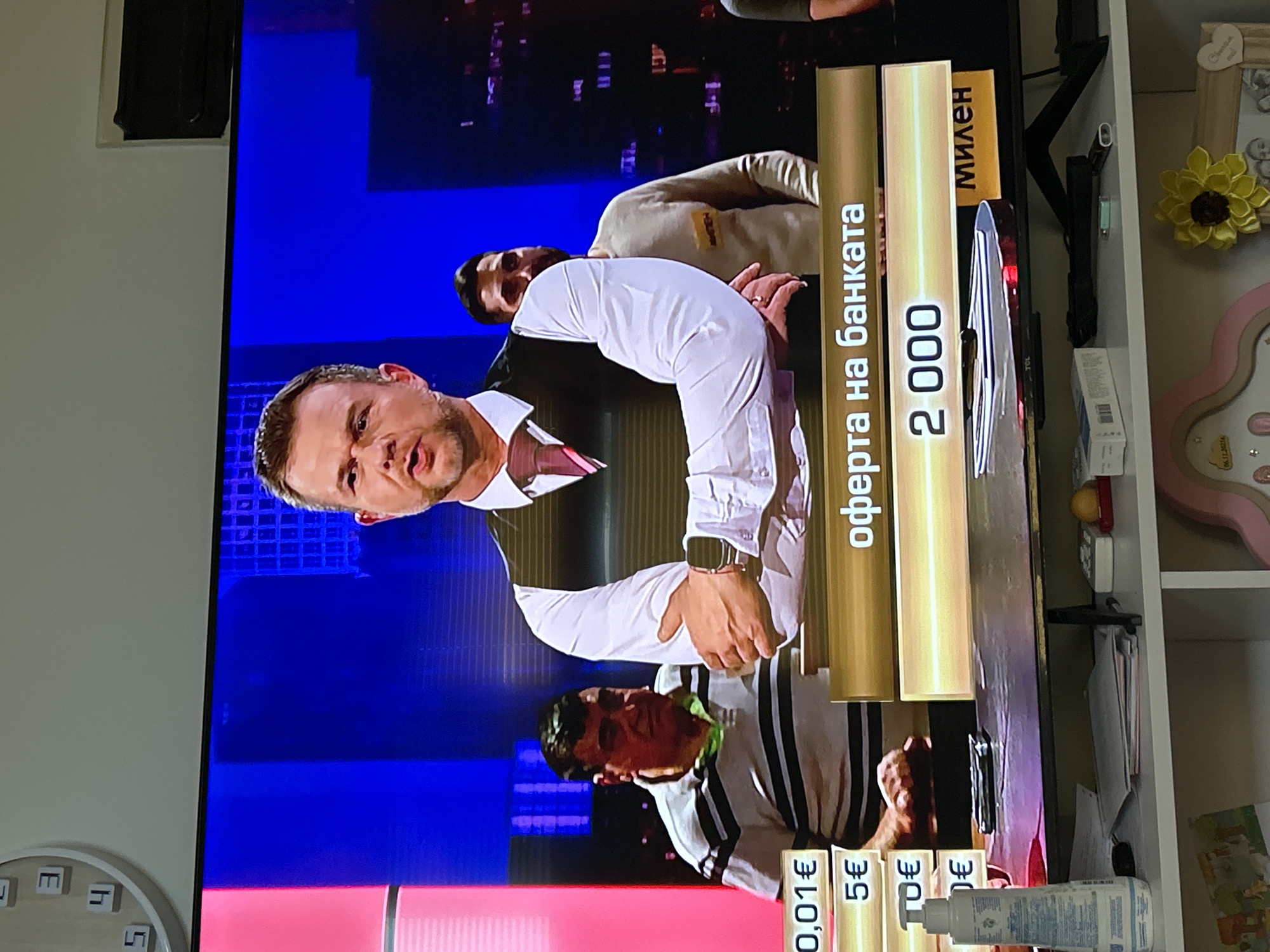 The image shows a television screen displaying a game show or similar program, captured from a slightly angled perspective and rotated 90 degrees clockwise.
On the television screen, three men are visible. The most prominent figure, centered, is a man with short brown hair and a beard, wearing a white collared shirt and a dark vest, looking intensely forward. Above him, another man in a light-colored sweatshirt with a "МИЛЕН" (Milen) name tag is partially seen. Below the central figure, a third man in a black and white striped shirt is visible from the chest up. The background on the TV is a bright blue, suggesting a studio setting.
Prominently displayed on the right side of the screen are two golden vertical bars. The left bar reads "оферта на банката" (offer from the bank) in Cyrillic, and the right bar shows the number "2 000". Along the bottom edge of the screen, several smaller vertical bars display monetary values in Euros: "0,01€", "5€", "10€", and another partial "€" amount.
The television itself is surrounded by a home environment. To the left, a plain light-colored wall is visible. To the right, a white shelf unit holds several decorative items, including a yellow fabric flower, a pink heart-shaped frame with the date "06.11.20258" inscribed, and a white bottle, possibly a lotion, with "Chantsue" visible on its label. A wall clock, partially visible on the bottom left wall, shows some Cyrillic letters like "LT" and "Ш". The visible Cyrillic text and Euro currency are consistent with the location context of Kavarna, Bulgaria.