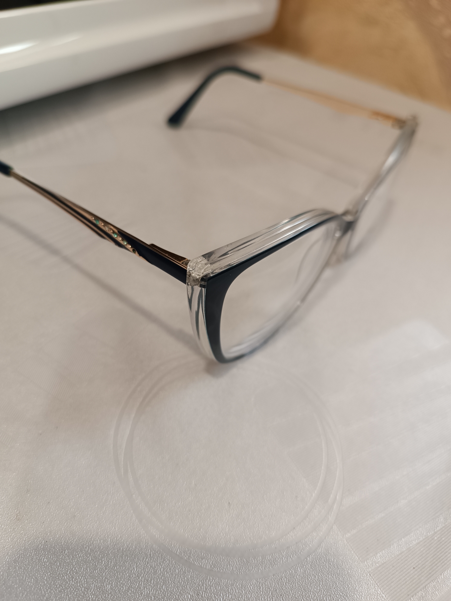 The image displays a pair of eyeglasses resting on a light-colored, textured surface. The eyeglasses feature a clear, transparent frame with a darker, likely navy or black, lower rim around the lenses. The arms (temples) of the glasses are made of a metallic, gold-toned material, accented with small, shimmering green details or gems along their length, particularly near the hinges. Parts of the arms near the front are also dark-colored, matching the lower lens rim. The lenses themselves are clear.
The surface beneath the glasses appears to be a placemat or tablecloth, white or off-white, with a subtle textured pattern. Visible on this surface are faint, circular imprints, possibly left by a glass or cup, and some linear or grid-like patterns, suggesting a plastic or woven material. In the background, a blurred, light-colored object, possibly a piece of furniture or an appliance, is visible. The scene is set indoors. No specific activity is occurring; the glasses are simply positioned on the surface. The time of day and weather are not discernible. There is no visible text in the image. Given the provided context, the image is from Chaikovsky, Russia, depicting a common household item in an ordinary indoor setting.
