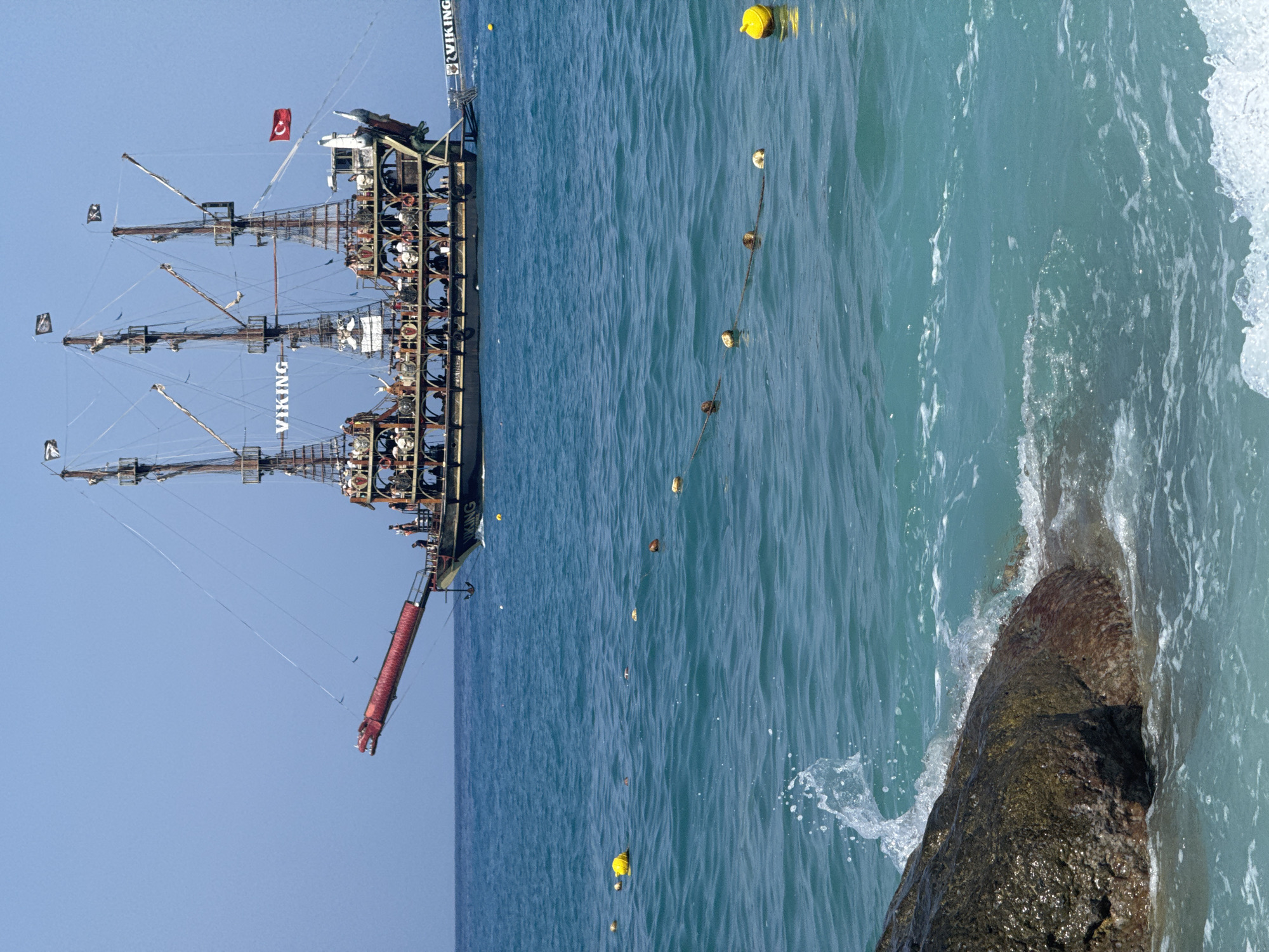 A large, multi-masted pirate-themed tour boat named "VIKING" is sailing on turquoise waters under a clear blue sky. The boat has several decks filled with passengers. Its bow is adorned with a red dragon's head figure. A Turkish flag is visible on the stern. In the foreground, waves crash against a large rock. Yellow buoys are spaced out in the water, attached to a rope. The scene is consistent with the coastal areas of Fethiye, Turkey, likely a popular tourist destination.
