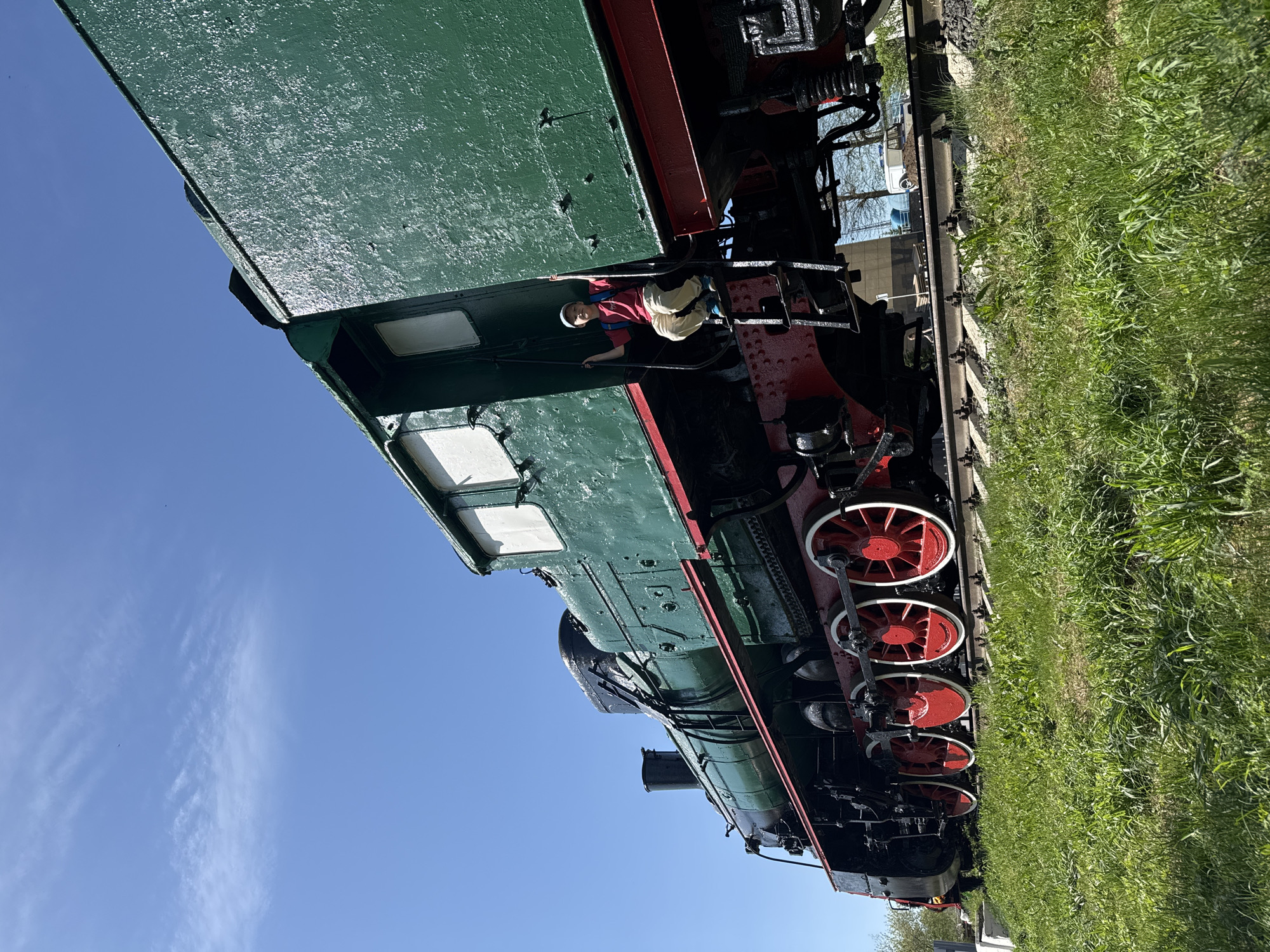 The image captures a green and red steam locomotive, likely a preserved exhibit, situated on train tracks amidst green grass. The sky is clear and blue. A young boy in a red shirt, light-colored pants, and a white hat is climbing on the train. The photograph was taken in Balezino, Russia.