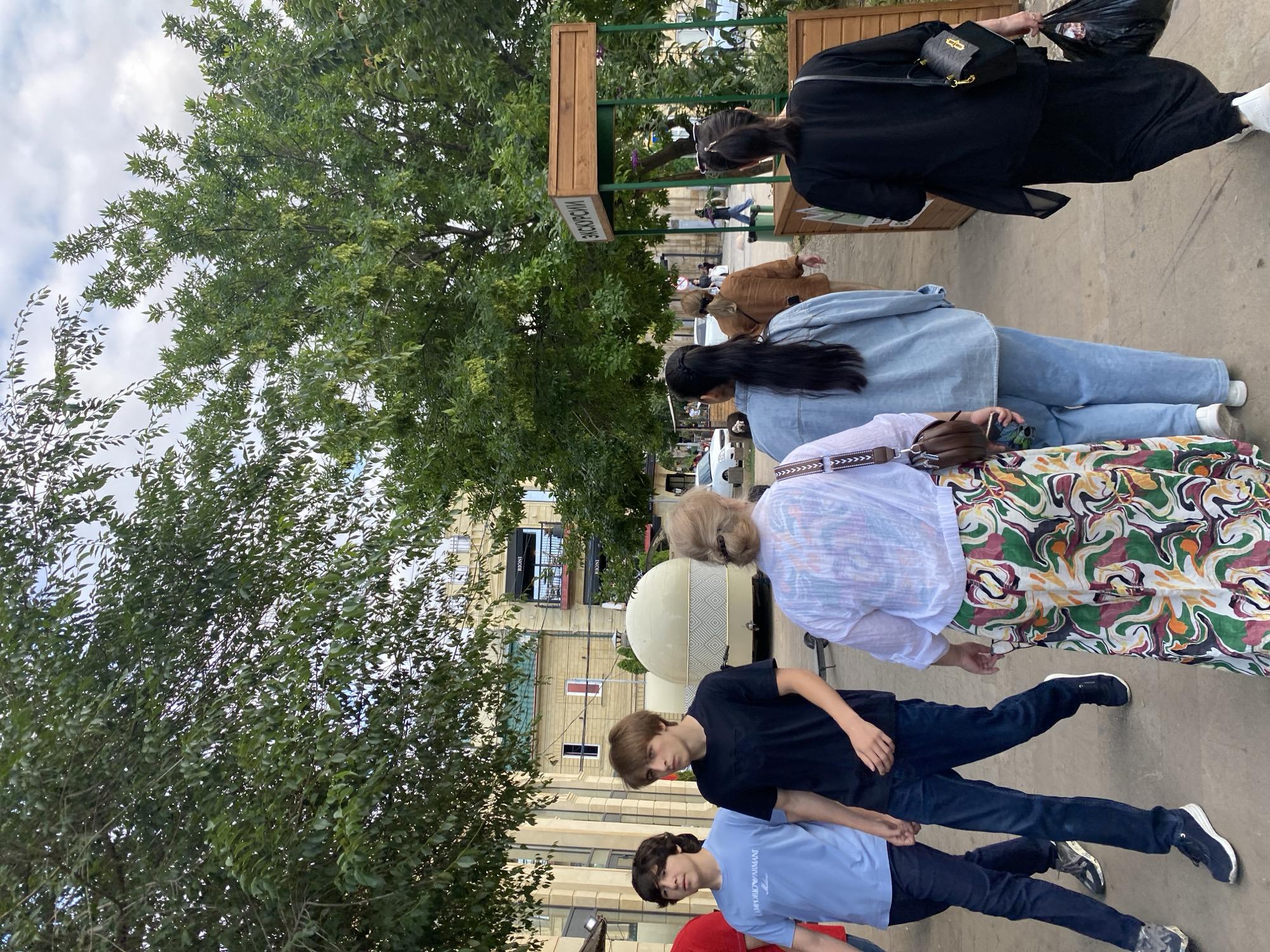 The image depicts a group of people walking on a paved pathway outdoors, with lush green trees and buildings in the background. Several individuals are walking in the same direction, and their attire suggests a casual outing.
In the foreground, two young males are walking side-by-side. The one closer to the camera is wearing a light blue t-shirt with "EMPORIO ARMANI" printed on it. The other young man is wearing a black t-shirt and dark blue jeans. Behind them, an older woman is visible from the back, wearing a patterned skirt and a white top. Further back, a person in a light blue denim jacket and jeans is walking, followed by another individual in a brown jacket and a person in a black outfit.
The scene appears to be in a public area, possibly a park or a pedestrian zone, given the presence of trees, benches, and architectural elements of buildings. The lighting suggests it is daytime, likely with overcast skies, as the light is diffused.
A wooden sign with Cyrillic text is partially visible on a green structure in the mid-ground, with the visible portion reading "ИЗБОРСКИЕ". This is likely a place name or a sign related to a business or attraction. The buildings in the background have a somewhat modern architectural style, with balconies and windows.
The overall mood of the image is neutral, capturing a moment of everyday activity with people on the move. No specific emotions are overtly expressed, but the activity suggests a leisurely stroll or perhaps commuting. The presence of trees and the general setting indicate a pleasant, possibly warm day. The context of Derbent, Russia, places this scene in a specific geographical and cultural setting.