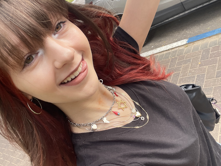 The image displays a young female captured in a high-angle selfie, smiling directly at the camera with an open mouth, revealing teeth and a slightly extended tongue. She has long, dark brown hair with significant red highlights and bangs, and light brown eyes accented with white eyeliner. She is wearing a black short-sleeved t-shirt and silver hoop earrings. Her neck is adorned with multiple layered necklaces: a thick silver chain featuring various charms including a heart, a map of Africa, a red tear-drop, a red chili pepper, and a silver figure, alongside gold-colored chains with a prominent Star of David pendant, a heart pendant, and another small Star of David. A faint reddish mark is visible on the right side of her neck. A black bag is partially visible on her left side. The background consists of an outdoor urban environment, specifically a paved sidewalk made of light brown rectangular pavers arranged in a herringbone pattern. Adjacent to the sidewalk is a curb, marked with a blue painted line indicating typical Israeli parking regulations. A dark vehicle or object is partially visible beyond the curb. The scene is located within the city of Raanana, Israel.