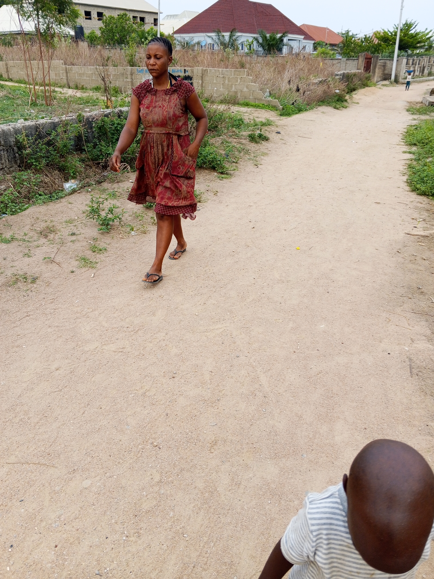 The image shows a woman walking on a dirt road in Adonkolo, Nigeria. She is wearing a reddish-brown patterned dress and sandals, and her hands are in her pockets. The road is sandy and dusty, with patches of grass and weeds along the sides. In the background, there are houses and concrete block walls. A child wearing a striped shirt is visible in the lower right corner of the frame, and another individual is seen farther down the road in the distance.