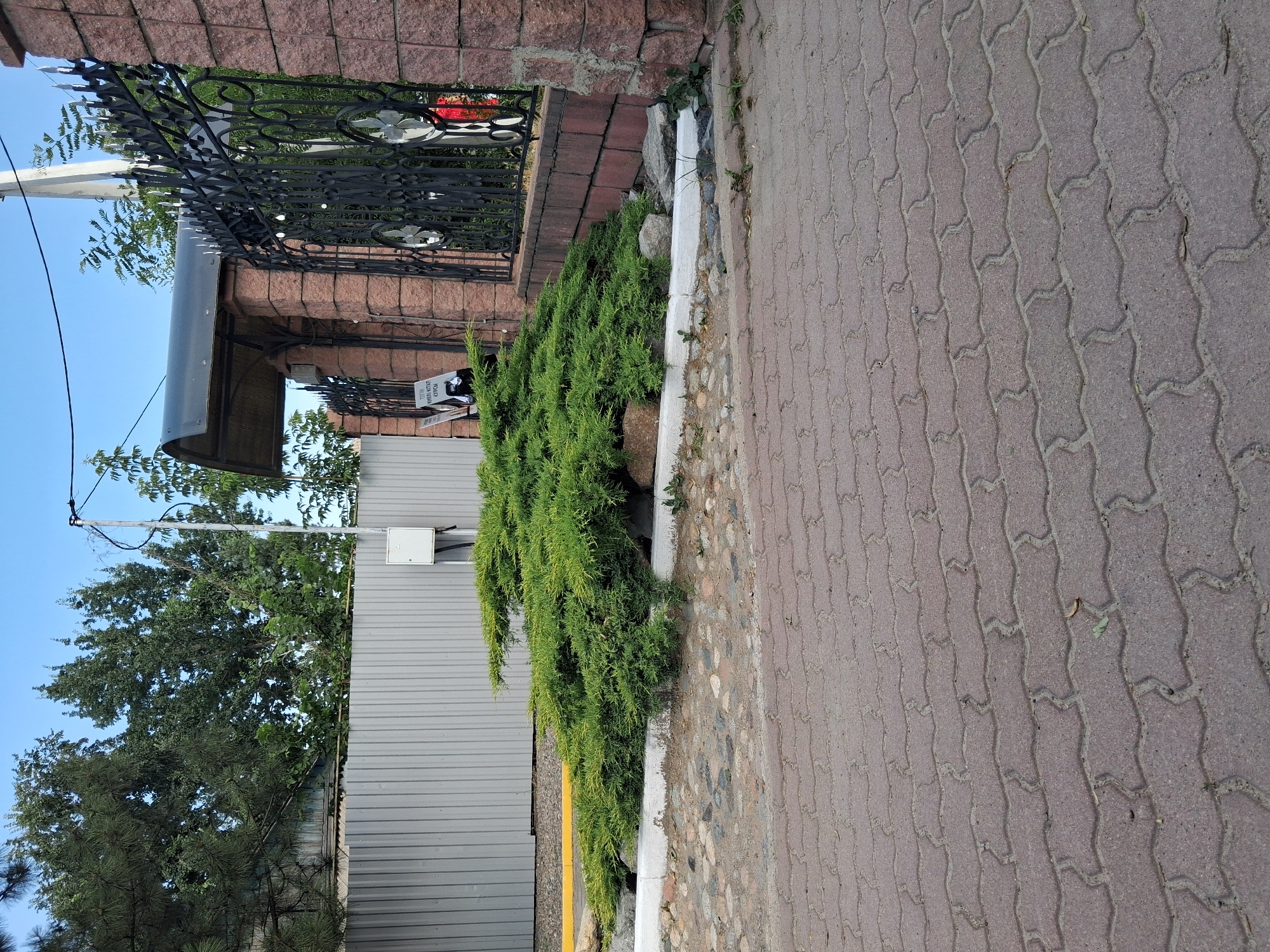The image depicts an outdoor scene, likely on a paved walkway or driveway made of interlocking red bricks. In the foreground, the brickwork is the dominant feature. Behind the bricks, there is a low, curved wall or border with a white coping stone. Adjacent to this is a patch of ground covered with small, irregular stones and some scattered green foliage, including a large, sprawling green shrub with yellowish-green tips. Further back, a tall, grey corrugated metal fence runs horizontally across the image. To the right of the metal fence, a section of a brick wall, constructed from reddish-brown stones, stands tall. This wall is topped with an ornate black metal fence featuring intricate scrollwork and finials. A dark, curved awning is visible above an entranceway between pillars adorned with the same reddish-brown stonework. A small white electrical box is mounted on the corrugated metal fence. The sky above is a clear, light blue. The scene appears to be an exterior view of a property, possibly a residential or commercial entrance, in the city of Baterk, Kazakhstan. There are no people visible in the image.