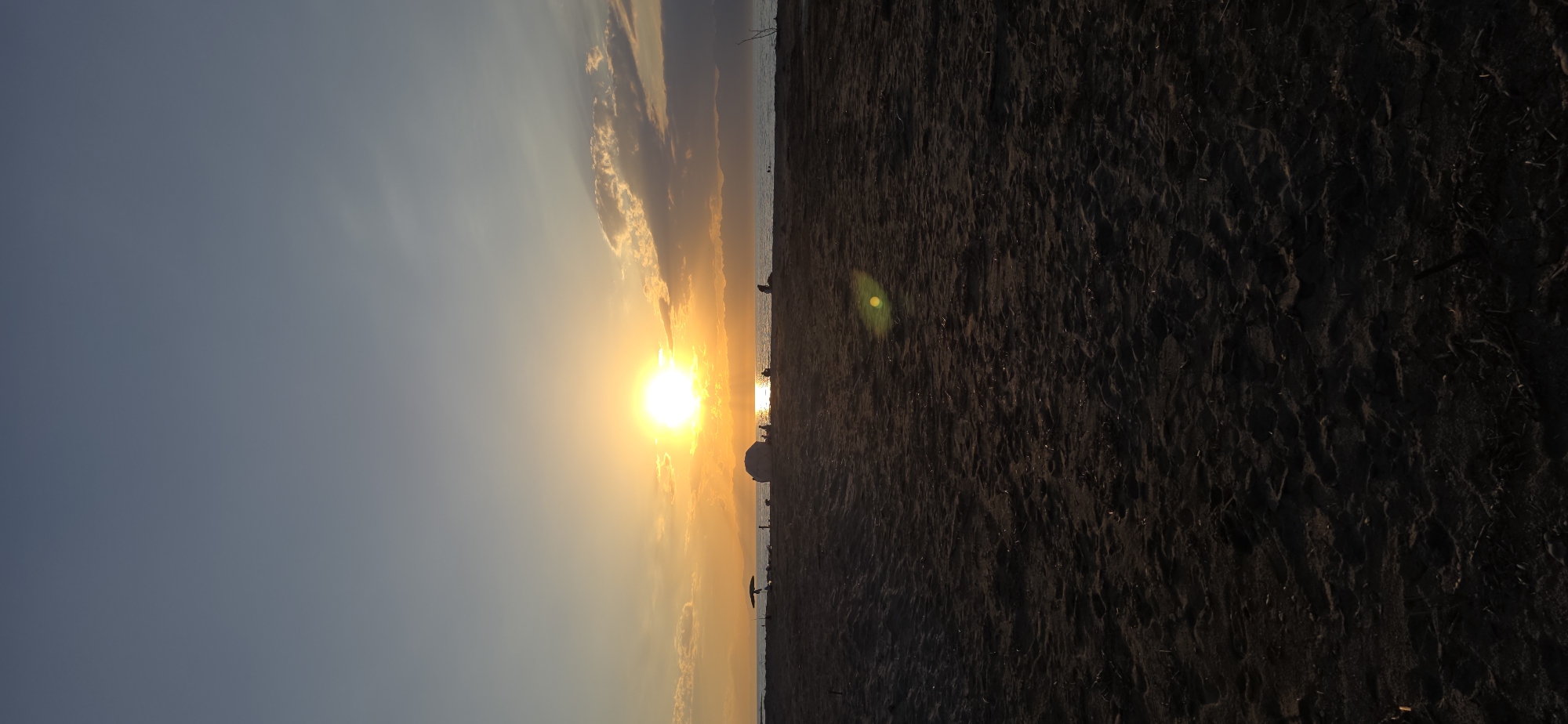The image depicts a serene sunset over a beach. The sun, a bright orb, is positioned just above the horizon, casting a warm, golden glow across the sky and reflecting on the water. The sky transitions from a deep blue at the top to hues of orange and yellow near the horizon. Scattered clouds are visible, catching the sunlight.
In the foreground, a sandy beach stretches across the frame. The sand appears dark and textured, with visible footprints and debris. In the middle ground, several figures are present on the beach, some sitting or standing, silhouetted against the bright sunset. An umbrella is visible to the left of the frame. A large, rounded object, possibly a rock or a large object, is situated on the beach near the water's edge. The water itself appears calm, with gentle waves lapping the shore. The distant horizon is marked by a range of mountains, silhouetted against the colorful sky. The overall scene suggests a peaceful end to the day, with people enjoying the natural beauty of the beach at sunset. The location provided, Boz Beshik, Kyrgyzstan, indicates a setting in Central Asia, known for its mountainous terrain and natural landscapes.