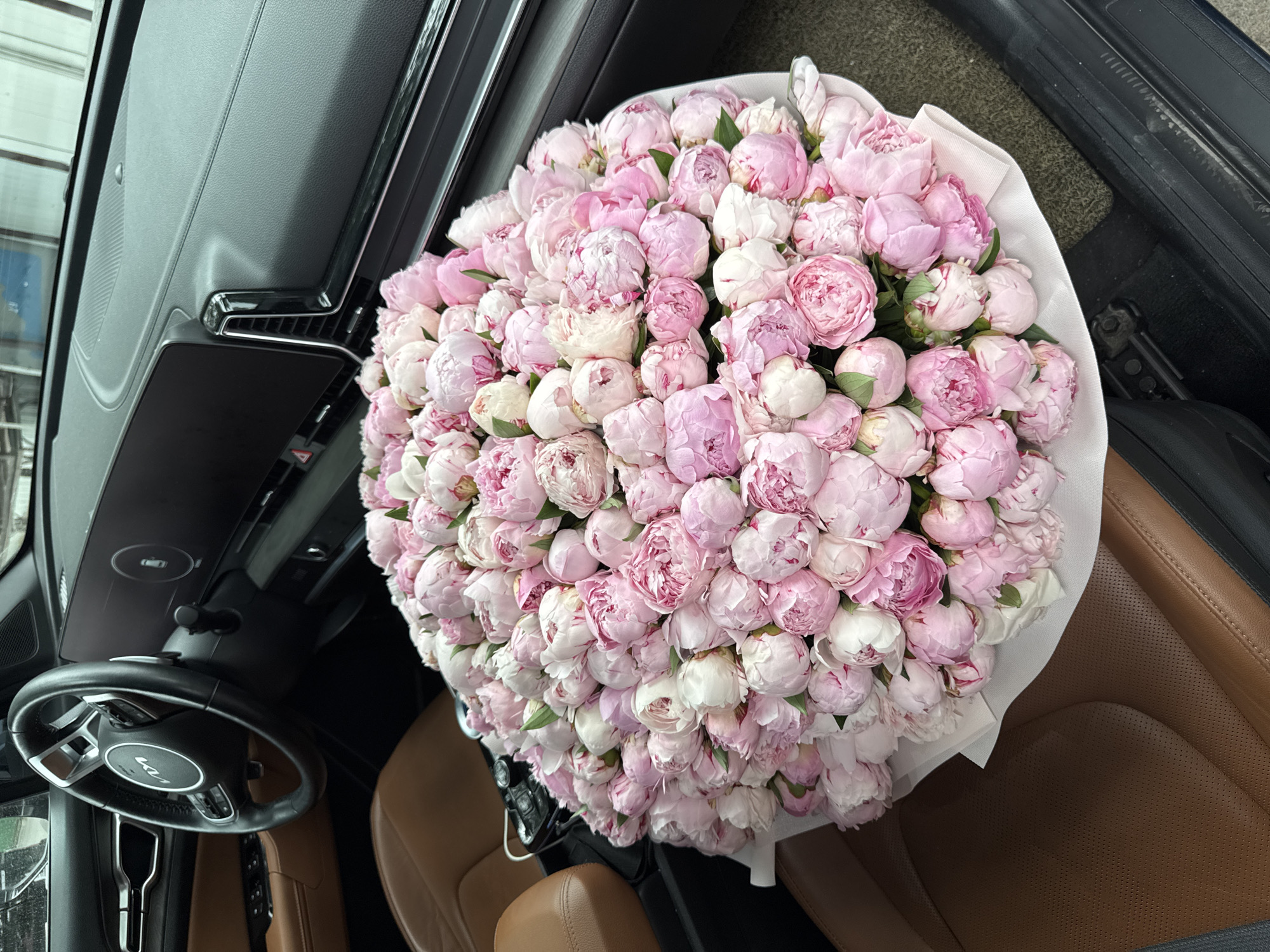 A bouquet of pink and white peonies is placed on the passenger seat of a car. The car interior features brown leather seats and a modern dashboard with a large screen and steering wheel. The background appears to be an urban environment, possibly Almaty, Kazakhstan, as suggested by the car's model and interior design, often seen in luxury vehicles within the city. No people are visible in the image.