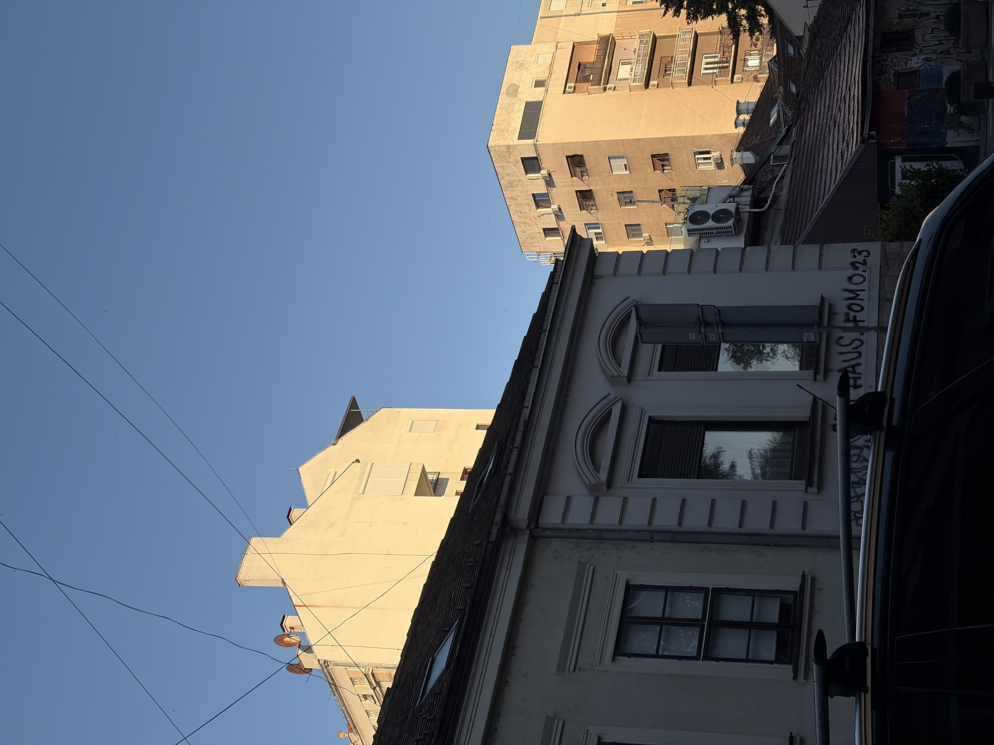 The image depicts a street scene in Belgrade, Serbia, under a clear blue sky. In the foreground, the roof rack of a dark vehicle is visible. Behind the vehicle, a low-rise building with a white facade features dark-framed windows and classical architectural elements. Further back, a taller, light-colored building with multiple windows and balconies rises, bathed in the warm glow of sunlight. To the right, another taller building with a tan facade and visible air conditioning units stands. The overall impression is a typical urban environment with older and newer structures. Graffiti is visible on the lower portion of the buildings.
