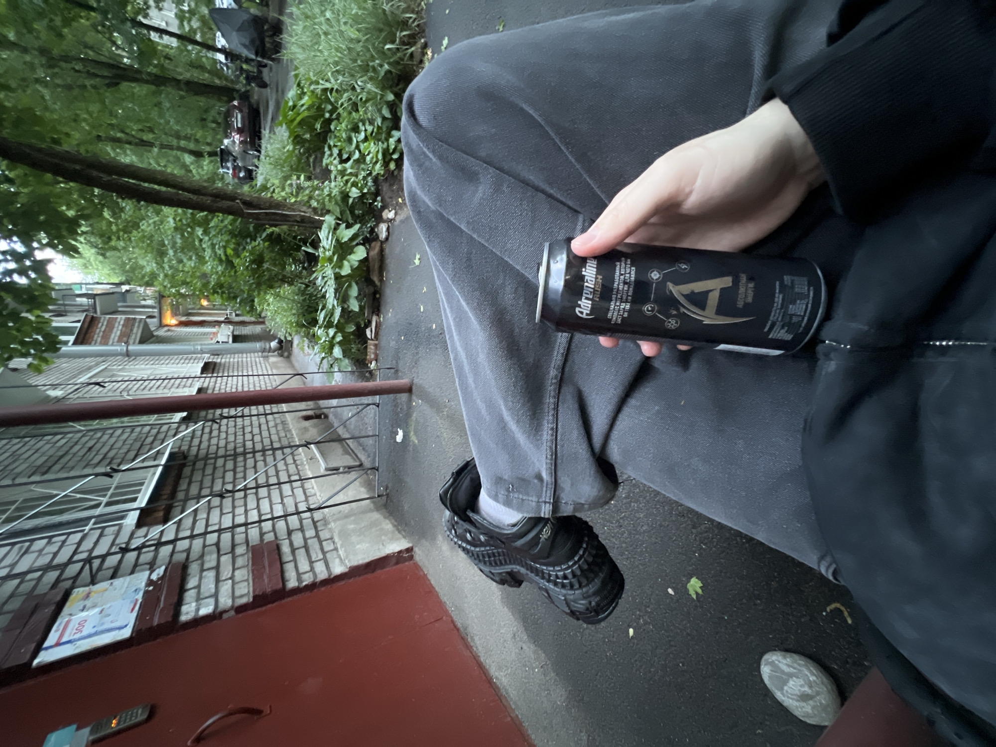 The image depicts a person sitting outdoors in Saint Petersburg, Russia, holding a can of "Adrenaline Rush" energy drink. The background shows a typical urban courtyard with brick apartment buildings, lush green vegetation, trees, and parked cars. The person is wearing dark grey pants and black athletic shoes. The environment appears wet, suggesting it may have recently rained. The focus of the image is the can held in the person's hand, with the logo and text clearly visible.