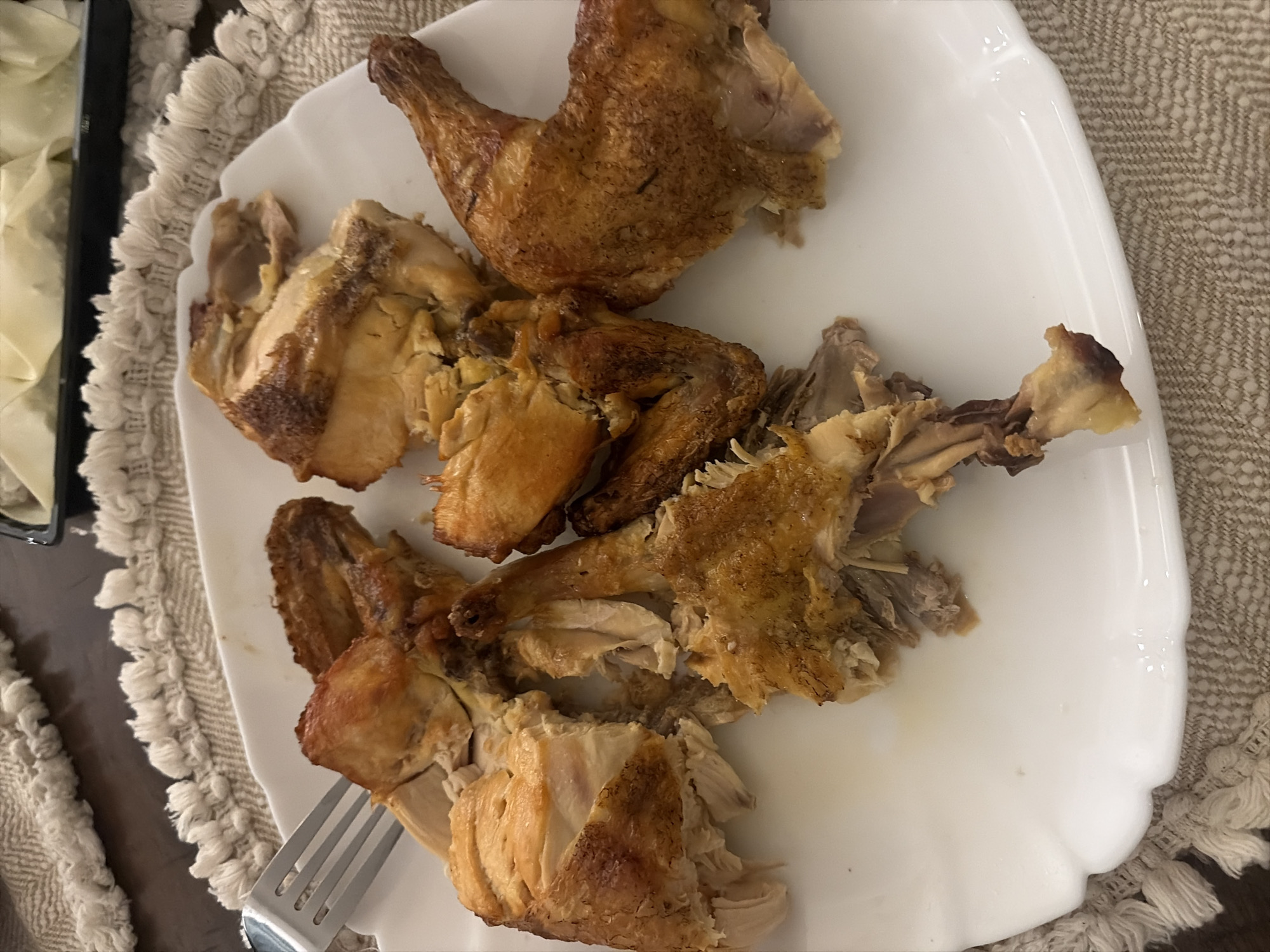 The image shows a white plate with pieces of cooked chicken. A silver fork is visible at the bottom left corner of the plate. The plate rests on a patterned surface, likely a tablecloth or placemat. Another dish is partially visible on the left side of the image, containing light-colored folded items. The environment appears to be a dining setting. There is no information available to suggest it is in Tbilisi, Georgia.