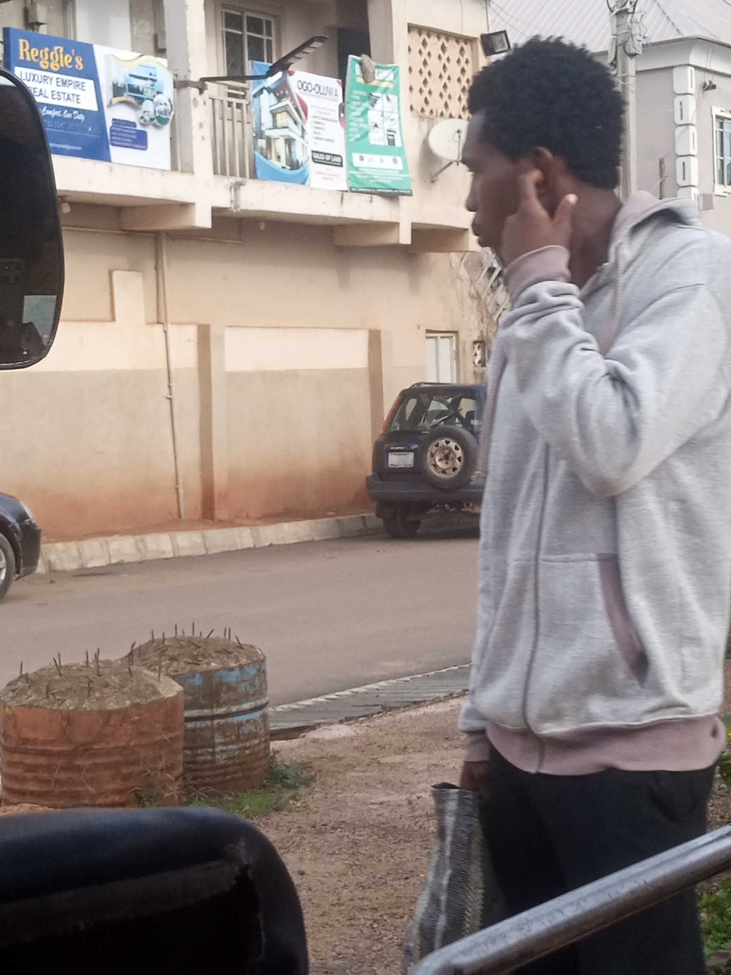 The image depicts a street scene in Rayfield, Nigeria. In the foreground, a young Black man, wearing a grey hoodie and black pants, stands with his right hand to his ear as if listening or speaking on a phone. He is also holding a patterned bag in his left hand. Behind him, a blue SUV with a spare tire mounted on the rear is parked on the street. Further back, several buildings with multiple stories are visible, some adorned with advertisements. One advertisement features the text "Reggie's LUXURY EMPIRE REAL ESTATE." Two large, rusted barrels filled with debris and topped with metal spikes are situated on the side of the road. The overall environment suggests an urban setting with ongoing development or construction indicated by the spiked barrels.