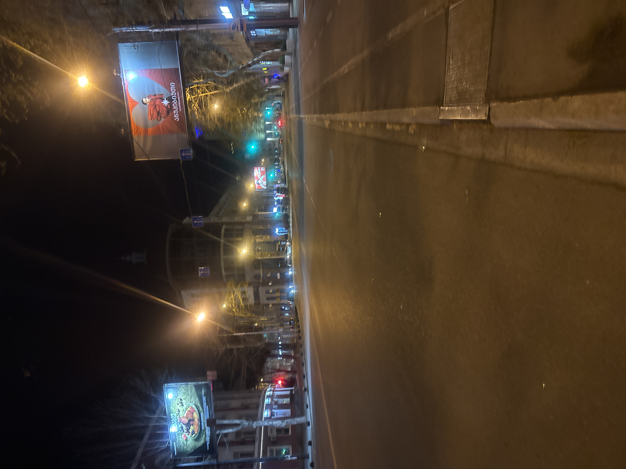 The image depicts a street scene at night in Tbilisi, Georgia. The view is from a low angle, focusing on the road that stretches into the distance, flanked by sidewalks and buildings on either side. The street appears to be largely deserted, with minimal vehicular or pedestrian traffic visible. Streetlights illuminate the scene, casting long shadows and creating a stark contrast between light and dark. Billboards with Georgian script and images are seen on the left side of the street. The architecture is a mix of modern and older buildings, indicative of the city's diverse architectural styles. The atmosphere is quiet and still.