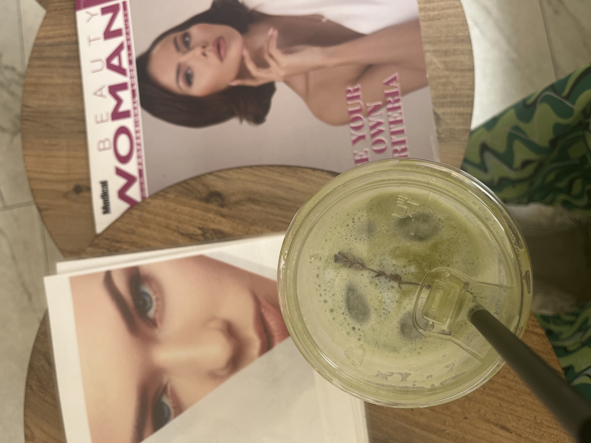 An overhead shot captures a close-up of a green beverage and magazines resting on a wooden table. In the foreground, a clear plastic cup with a lid contains a vibrant green drink, likely a matcha latte, topped with foam. A small sprig of a purple-flowered herb, possibly lavender, floats on the foamy surface. A black straw extends from the lid, which also bears a visible recycling symbol.
To the left of the drink, two magazines are partially visible. The top magazine is titled "BEAUTY WOMAN," with "Medical" printed in a smaller font beneath "BEAUTY." Its cover features a woman with dark hair, looking towards the viewer, with one hand elegantly poised near her face. Partially visible text on the cover reads "E YOUR OWN RITERIA," suggesting "DEFINE YOUR OWN CRITERIA." Below this, another magazine is seen, showing a partial view of a woman's face, specifically her eye, nose, and mouth, with fair skin and dark eyes.
The objects rest on a wooden surface with a distinct grain. In the upper right corner, a glimpse of a green, wavy-patterned fabric, possibly clothing or upholstery, is visible. The scene evokes a casual, relaxed atmosphere, perhaps in a café, waiting area, or home setting.