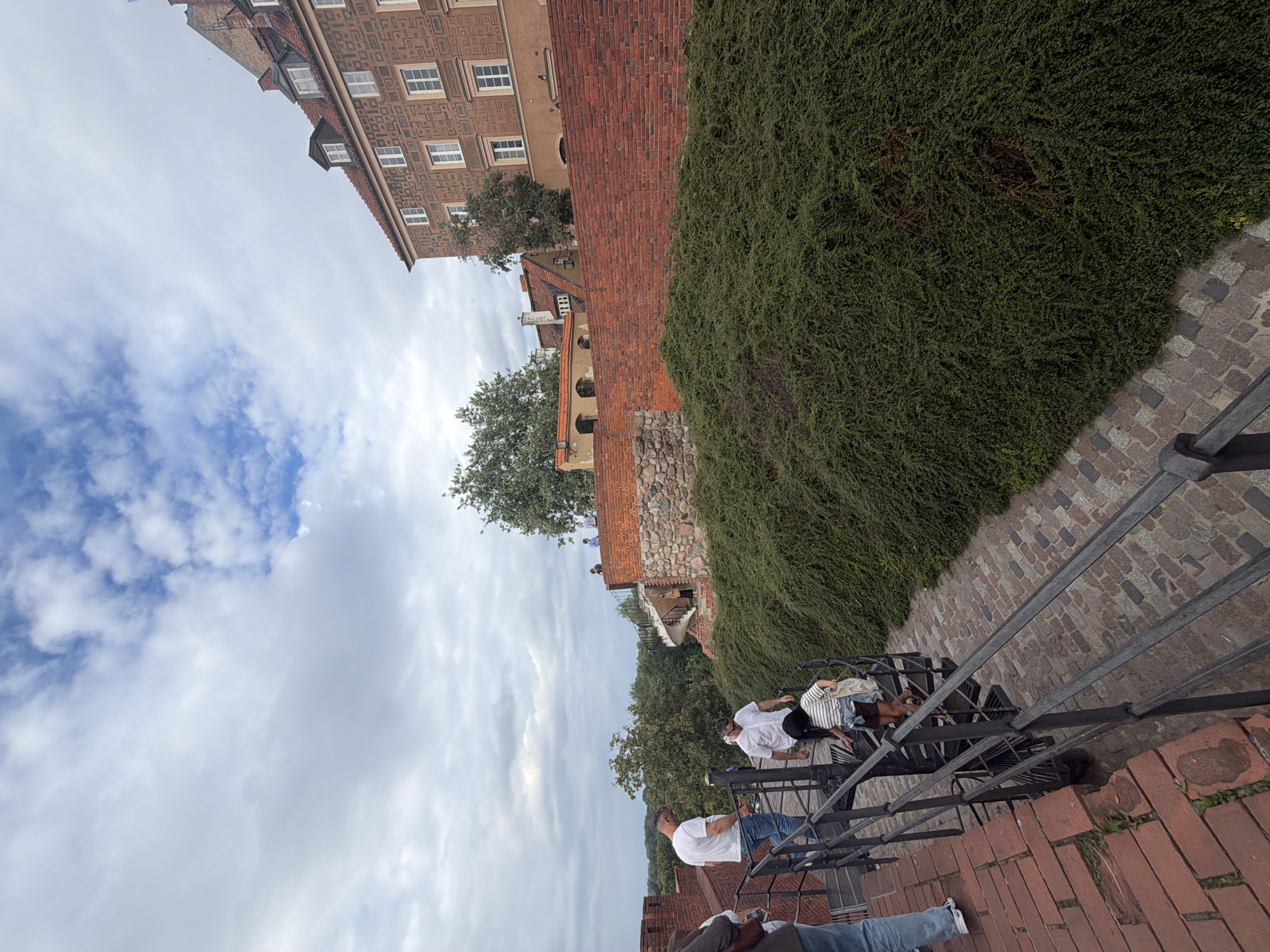 The image depicts an outdoor scene with several people on a tiered walkway. In the foreground, a person wearing blue jeans and a brown shirt is visible from the waist down, with their legs crossed. Further up the walkway, another person in a white shirt and blue jeans is standing. Above them, a group of individuals are gathered on a staircase, with one person in a white shirt and striped skirt, another in a white shirt and dark skirt, and a third in a white shirt.
The setting includes a brick building with numerous windows on the left side of the frame, appearing to be historical architecture. A thick, green hedge runs along the right side of the walkway, creating a natural barrier. The walkway itself is paved with cobblestones, and a dark metal railing runs along the edge, forming a staircase that leads upwards.
The time of day appears to be overcast, with a mix of clouds and patches of blue sky. The overall mood seems casual, with people enjoying the outdoors and the view. The architecture suggests a potentially historic or tourist location. No visible text is present in the image.