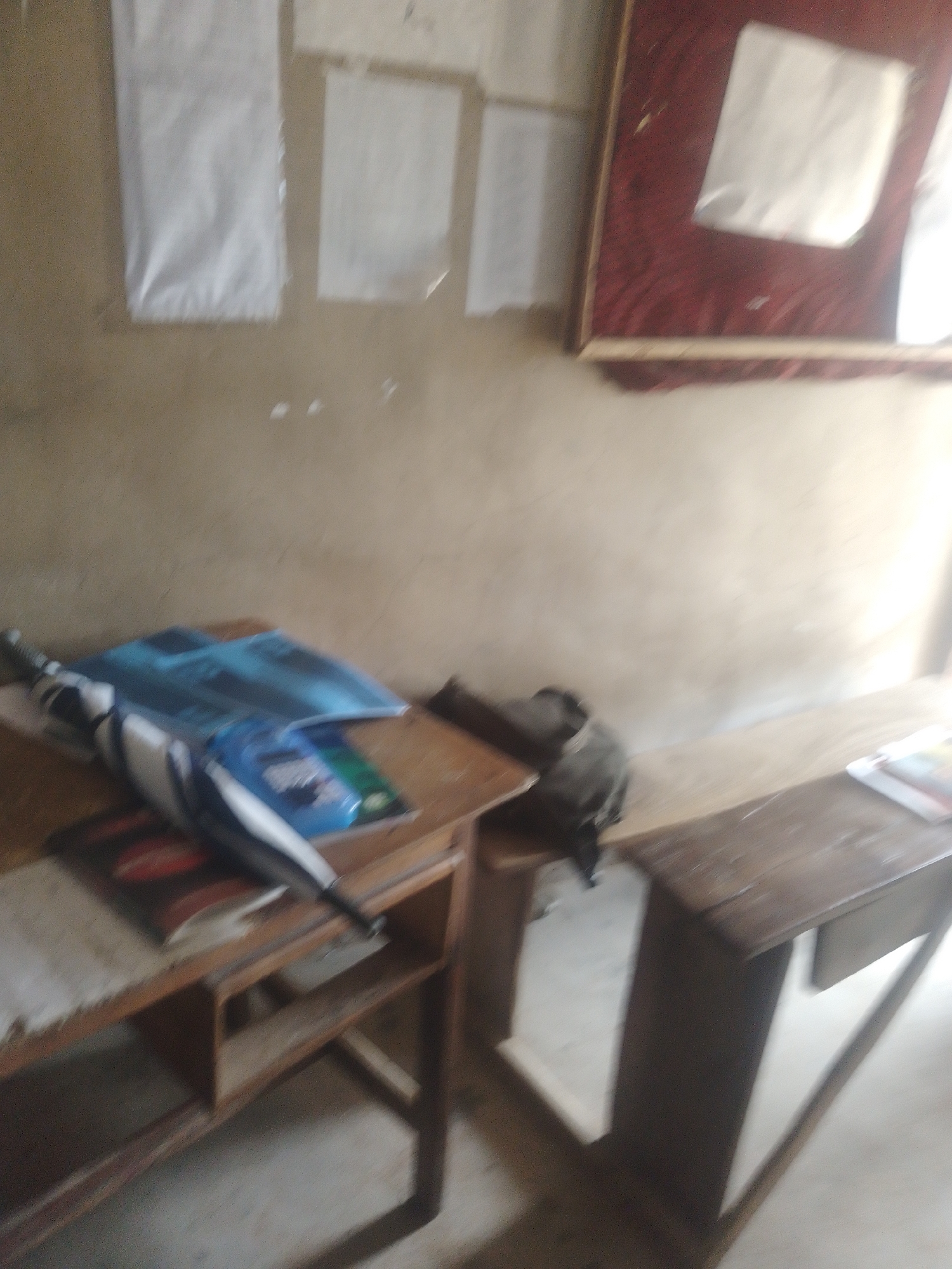 The image depicts an interior scene, possibly a classroom or study area, with elements suggestive of a rustic or simple setting. The walls are a muted beige color, with several sheets of white paper pinned to the upper portion. To the right, a large framed display with a dark red fabric background contains two white squares, possibly photographs or documents. In the foreground, a wooden desk is visible, cluttered with items. A blue object, possibly a folder or book, lies open on the desk. Partially covering it is a closed umbrella, its handle and fabric visible. To the right of the desk, a wooden bench runs along the wall. On the bench, a dark backpack or bag rests near the desk. Further to the right, another part of the bench holds a few items, including what appears to be a magazine or book. The lighting is uneven, with a bright light source on the right side of the image, casting shadows. The overall impression is one of a functional space that is perhaps a bit disorganized.