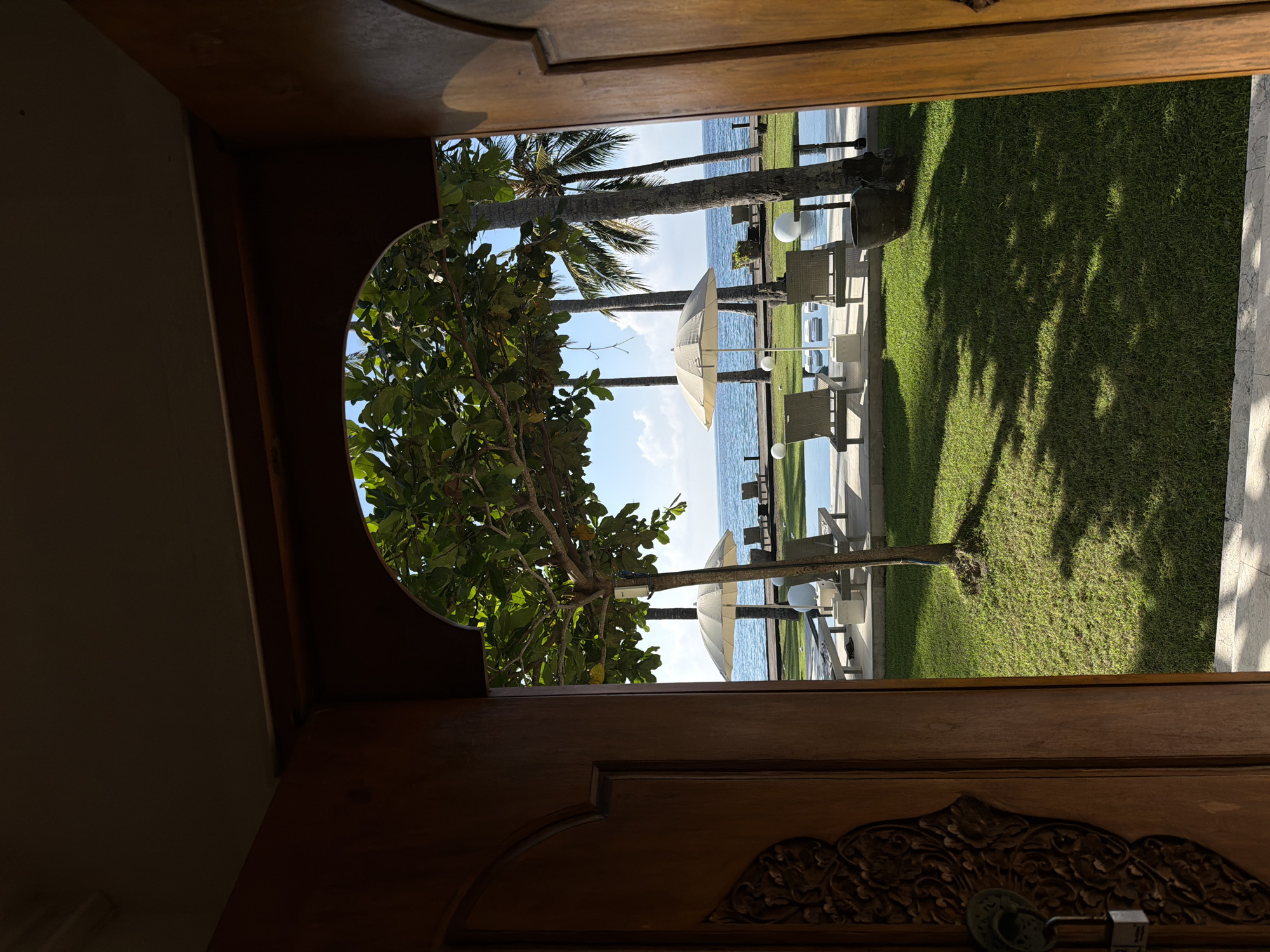 The photograph shows a view from inside a wooden door frame, looking out towards a tropical beach. The outdoor scene includes palm trees, a lawn, and beach umbrellas with chairs facing the ocean. The lawn appears green and well-maintained. The sky is blue. The location is likely in Karangasem, Indonesia.