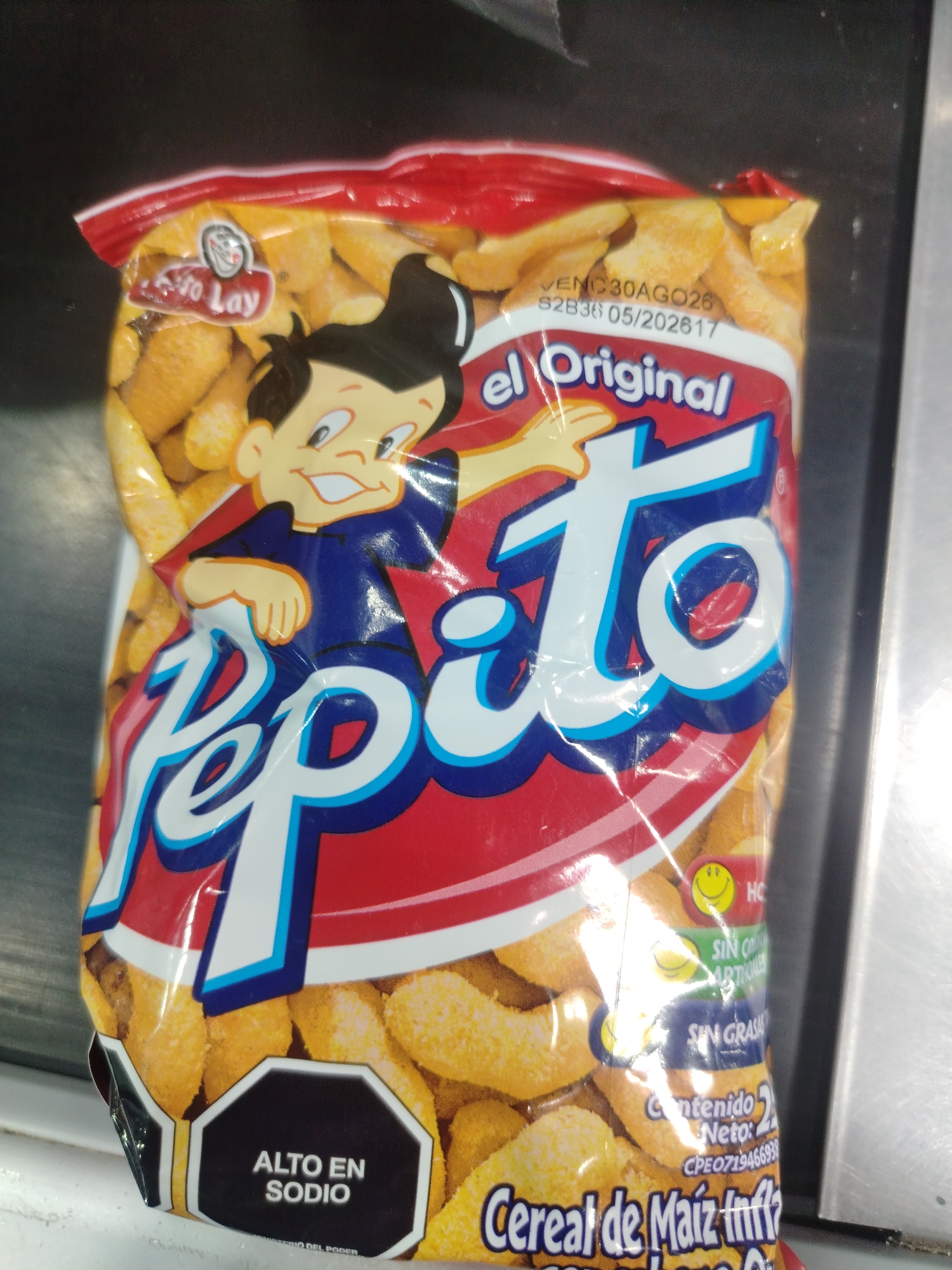 The image shows a bag of "Pepito" brand corn cereal snacks. The bag is red and yellow with a cartoon character and large white text that reads "Pepito". There is also text that says "el Original" and "Alto en Sodio". The bag is partially open, revealing the puffed corn snacks inside. The background appears to be a dark, reflective surface, possibly a countertop. The scene is likely indoors, in a commercial setting, as suggested by the clean, manufactured packaging and the reflective surface which could be part of a display or storage unit in a store. The location in Juan Griego, Venezuela is not discernible from the image itself.
