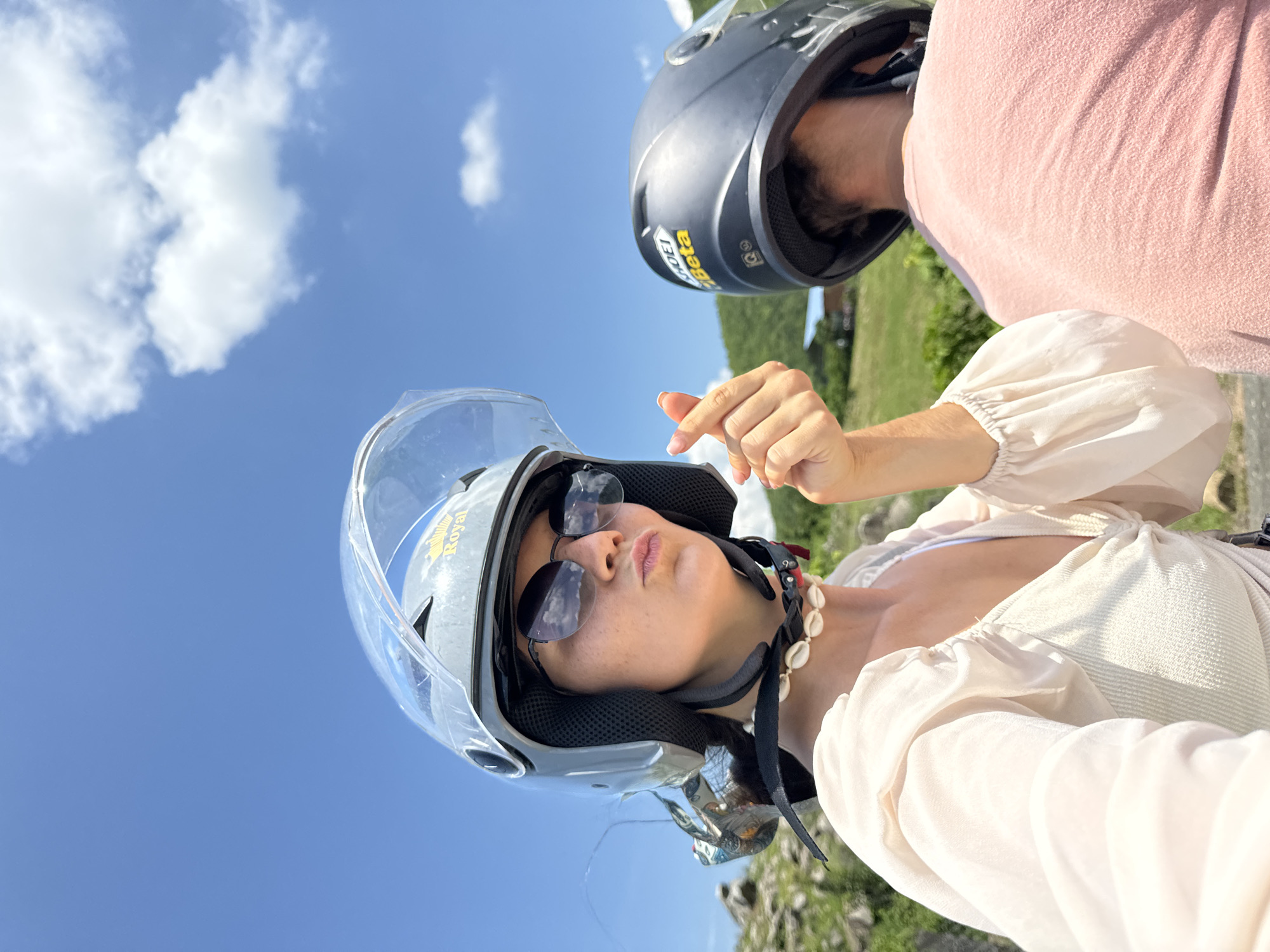 Two individuals, presumably on a motorbike, are captured in a selfie taken outdoors under a bright blue sky with scattered white clouds. The person in the foreground, wearing a grey helmet with a clear visor and sunglasses, is looking at the camera with their lips pursed and making a gesture with their hand that signifies "I love you" in Korean culture. They are also wearing a light-colored top and a seashell necklace. Behind them, partially visible, is another person wearing a dark helmet and a pink shirt. The background features lush green hills and some buildings, indicative of a rural or semi-rural setting. The presence of the helmets suggests they are traveling by motorbike, a common mode of transportation in Vietnam. The overall scene appears to be a casual moment captured during an outing, likely in a scenic location such as Hàm Thuận Bắc, Vietnam, given the terrain and vegetation.