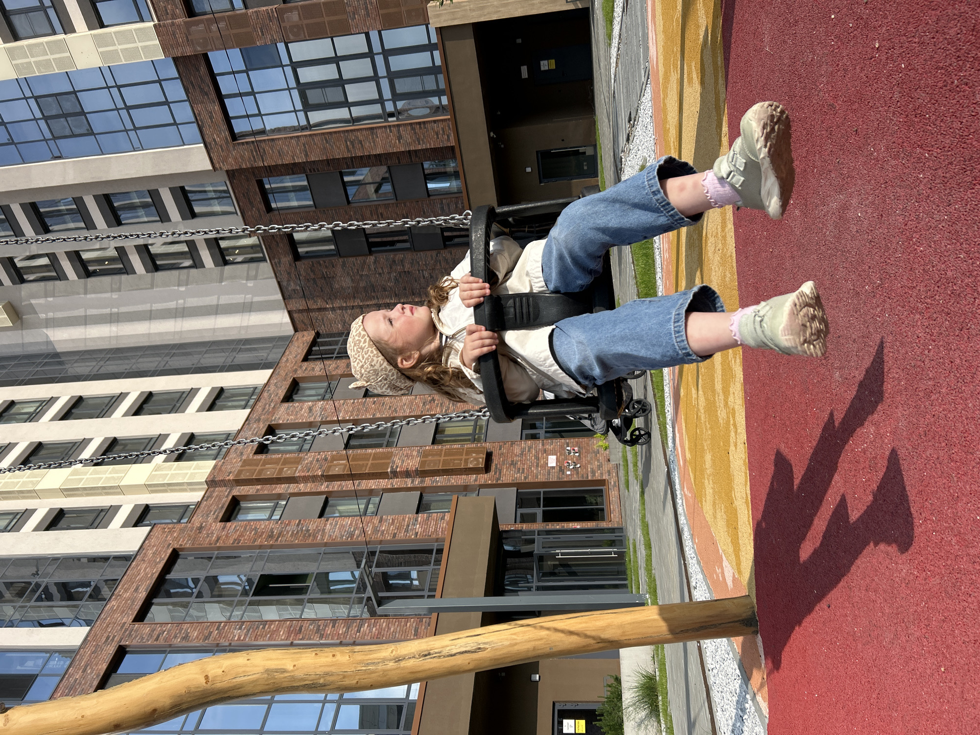 An outdoor, eye-level, medium shot captures a young girl with blonde hair and light skin, wearing a leopard print beret, a cream-colored jacket, and blue jeans, sitting on a swing. The girl is looking to her right with a slight smile. Her feet are in the air, wearing pink socks and light green sneakers. The swing set is made of dark metal and chains, with a thick, curved wooden pole on the left side of the frame. The background features a modern, multi-story building with a brick facade and large windows. The ground is covered with a red and yellow patterned rubber surface, with some gravel and grass visible. In the middle ground, a stroller is parked. The image appears to have been taken on a sunny day, casting shadows on the ground. The scene is a playground area within an urban setting, likely a courtyard of a residential complex, possibly in Yekaterinburg, Russia, given the architectural style of the building.