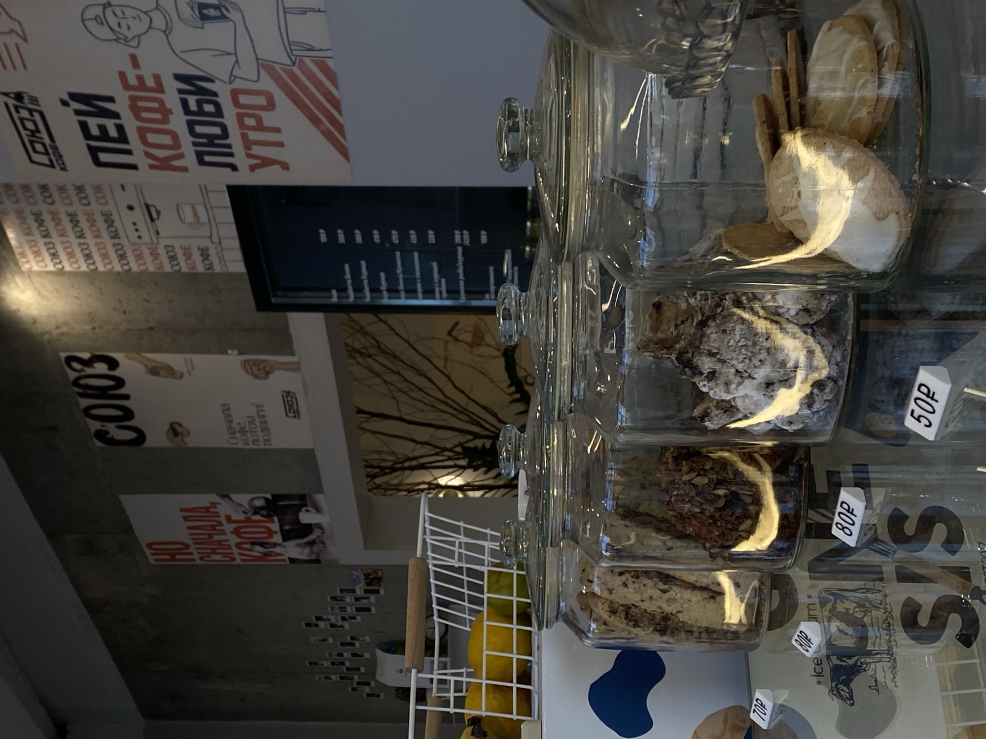 The image shows the interior of a cafe, likely in Agronom, Russia, as indicated by the Cyrillic text on posters. In the foreground, there are several large glass jars filled with cookies and other baked goods. Small white price tags with Cyrillic lettering are placed in front of the jars. The prices visible are "70P", "80P", and "50P". To the left, there is a white wire basket containing yellow fruits, possibly lemons or oranges. Behind the jars, there is a reflective surface that appears to be a display case or counter, with reflections of the baked goods and the environment. Further back, there are posters on a concrete wall. One poster features the text "НО СНАЧАЛА, КАФЕ" (But first, coffee) with an image of a dog. Another poster has "СОЮЗ" (Union) at the top and "ПЕЙ КОФЕ- ЛЮБИ УТРО" (Drink coffee - Love morning) with a drawing of a person holding a book. A black menu board is also visible behind the jars, displaying prices and item names in Russian. The overall lighting suggests an indoor setting, possibly with natural light coming from the left.