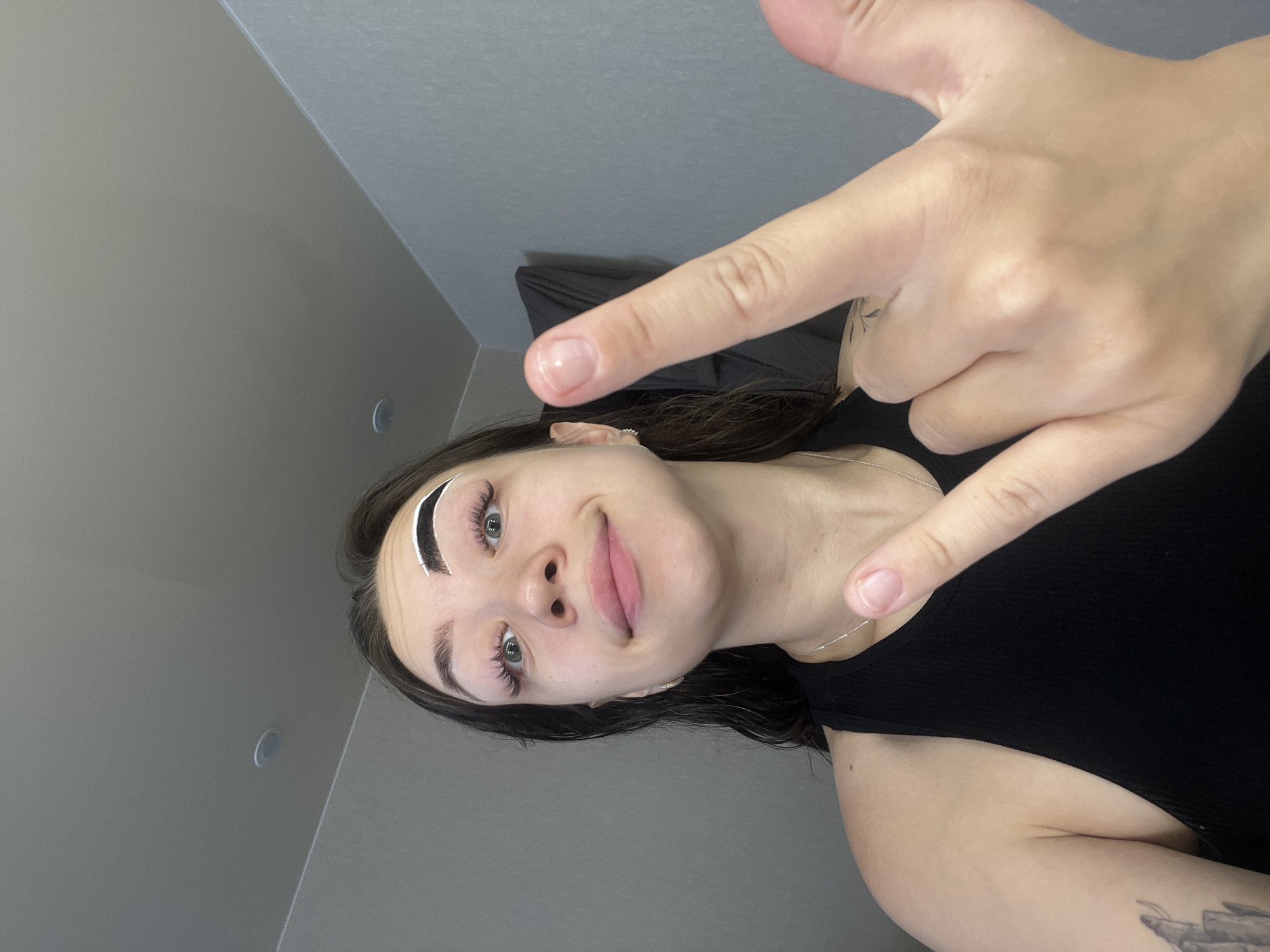 The image shows a young woman with dark hair and light skin, looking directly at the camera. She has heavy false eyelashes and is wearing a black tank top. Her right eyebrow has a white substance applied to it, possibly for shaping or tinting. She is holding up her right hand, with her index and middle fingers extended in a "rock on" gesture. Her left hand is not fully visible. The background appears to be an interior room with gray walls and a ceiling with recessed lighting. The location of Chaikovsky, Russia is not discernible from the image.