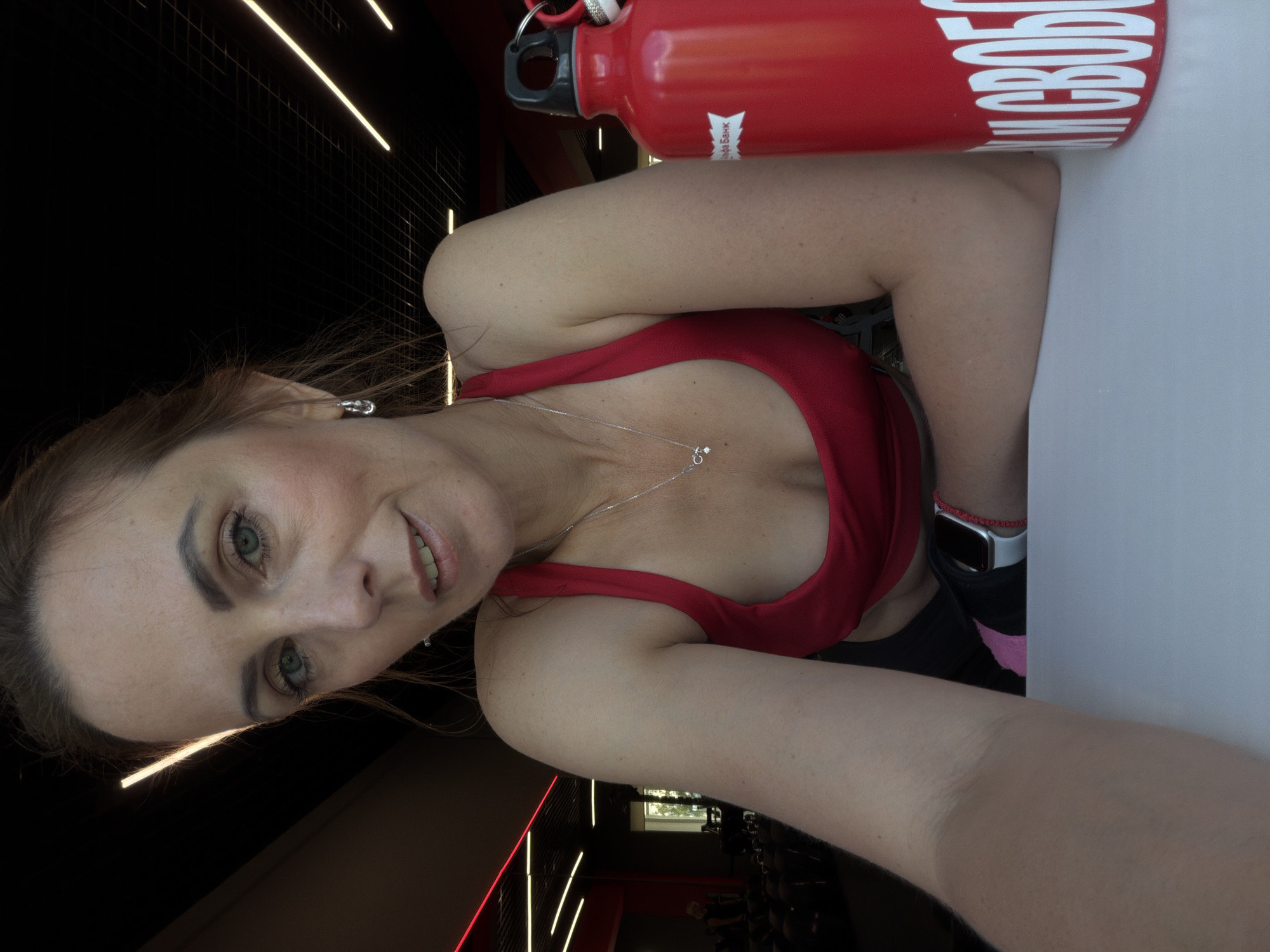 A woman is taking a selfie in a gym. She has blonde hair and is wearing a red sports bra. She is also wearing a silver necklace and a smartwatch on her left wrist. In the background, there is a red water bottle with white lettering that says "В СВОБОДЕ" (In Freedom) and a white logo. The gym appears to have a black ceiling with white lights and a dark tiled wall. The time of day is not specified, but the lighting suggests it could be daytime or evening. The woman's facial expression is a slight smile, indicating a positive emotion. The location context is Birobidzhan, Russia.