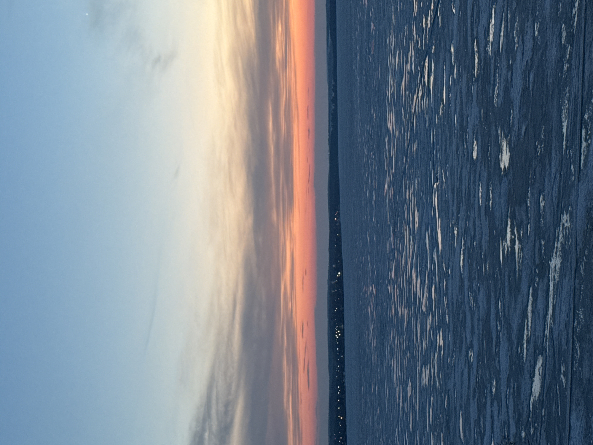 The image depicts a sunset over a body of water, likely a reservoir or lake, with a shoreline visible in the distance. The sky transitions from blue to orange and yellow hues, indicating the time of day. Reflections of light are visible on the water's surface. A line of lights on the shoreline suggests a settlement or urban area. The environment is natural, with atmospheric conditions influencing the light and color. Without additional information, it is difficult to confirm that the location is specifically in Votkinsk, Russia, but the scene aligns with its geographical context.