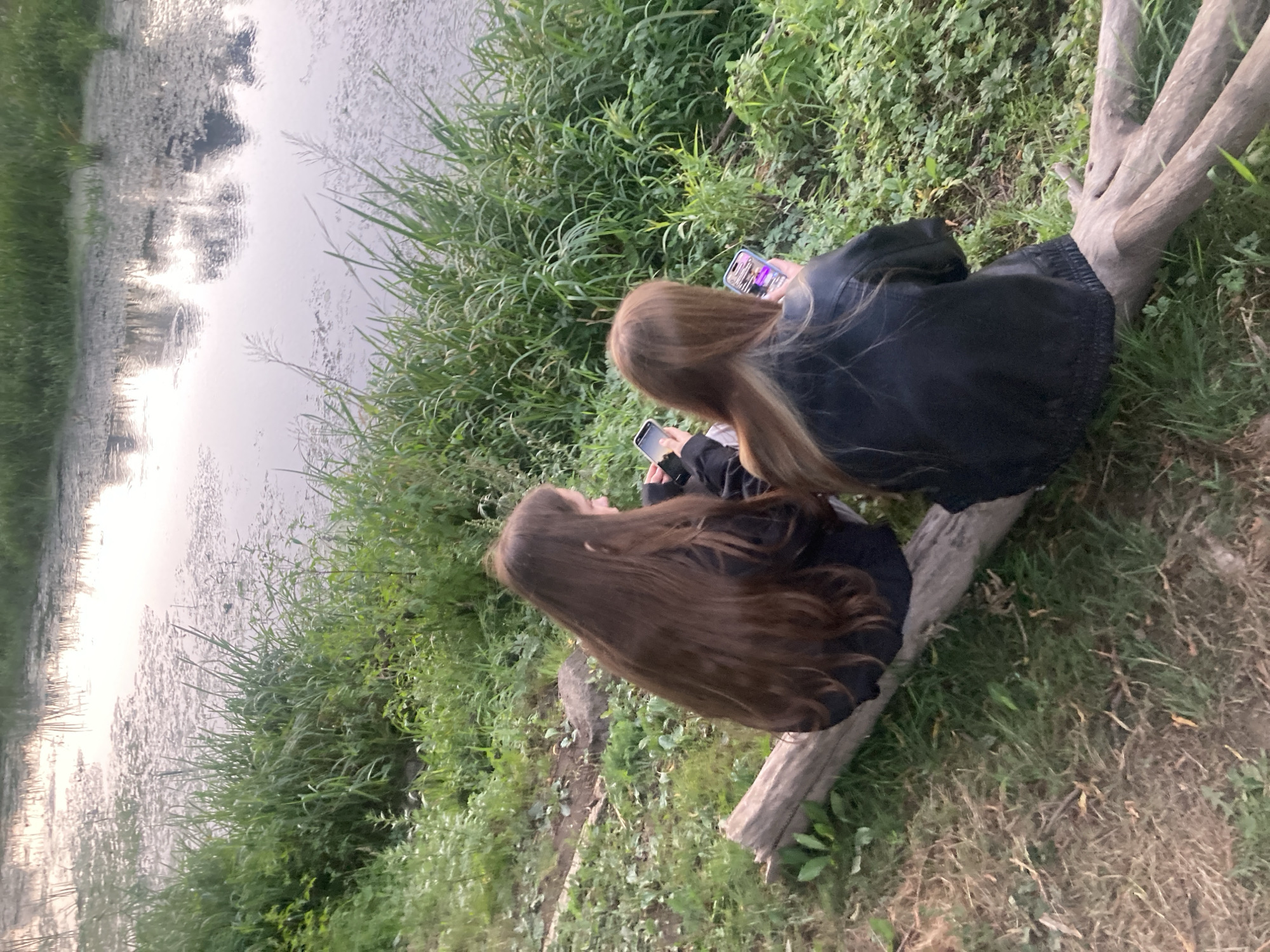 In the city of Brest, Belarus, two young individuals are seated on a fallen log by the water's edge. They are both looking at their mobile phones, with one person holding a phone displaying a colorful image. The environment is lush and green, with tall grass and other vegetation surrounding the water. The water itself appears calm, with reflections of the sky and surrounding trees visible on its surface. The overall scene suggests a peaceful and relaxed moment in a natural setting.