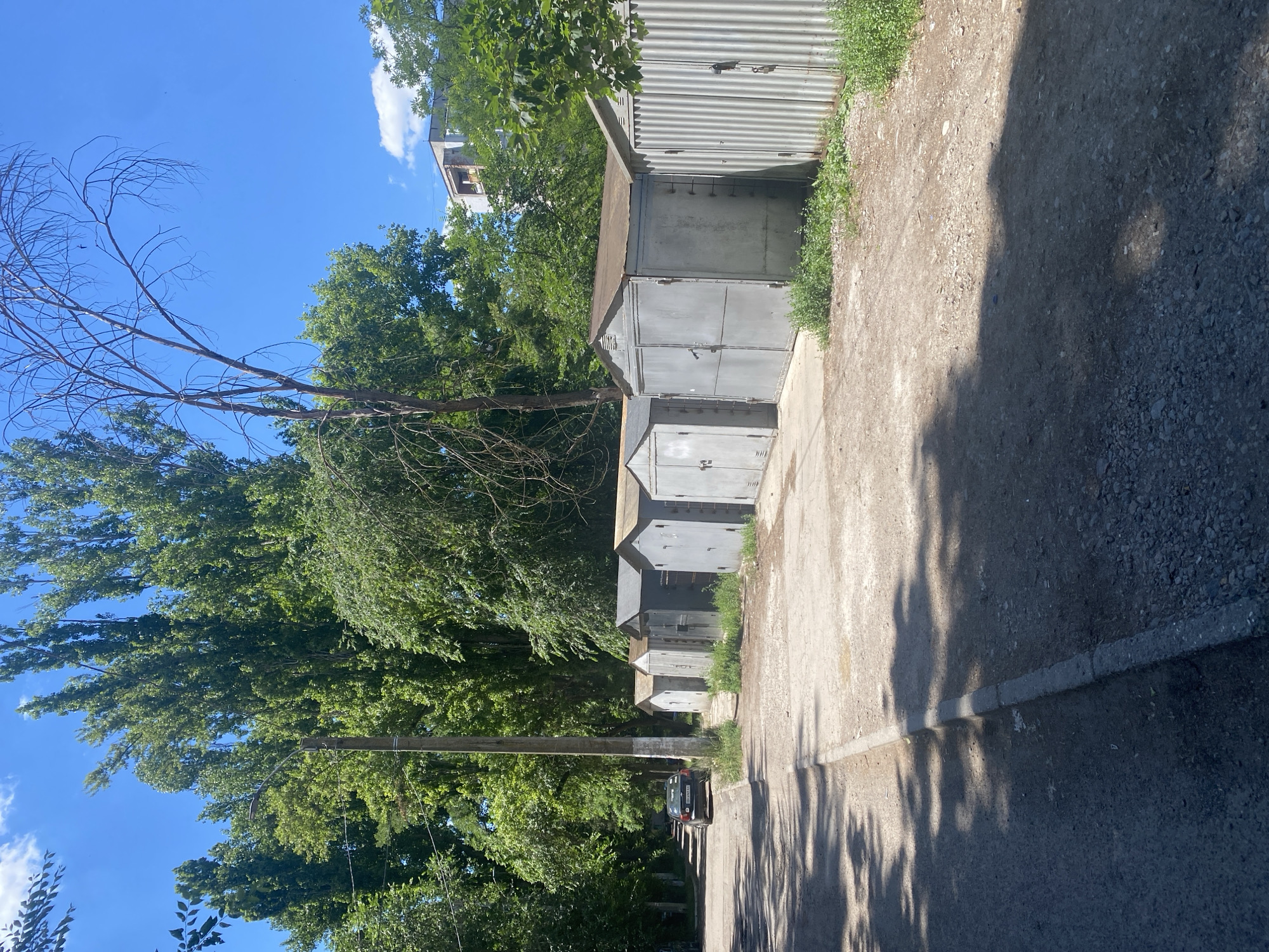 The image captures a row of grey metal garages situated in an urban setting, likely in Odesa, Ukraine, as suggested by the presence of a car with Ukrainian license plates. The garages are aligned along a dirt and gravel path, with a paved road and sidewalk visible to the left. Lush green trees line the background, casting shadows on the ground. A utility pole with a street lamp stands prominently near the garages. In the distance, a multi-story building is partially visible above the trees. The sky is a clear blue with a few scattered clouds. There are no people visible in the image.