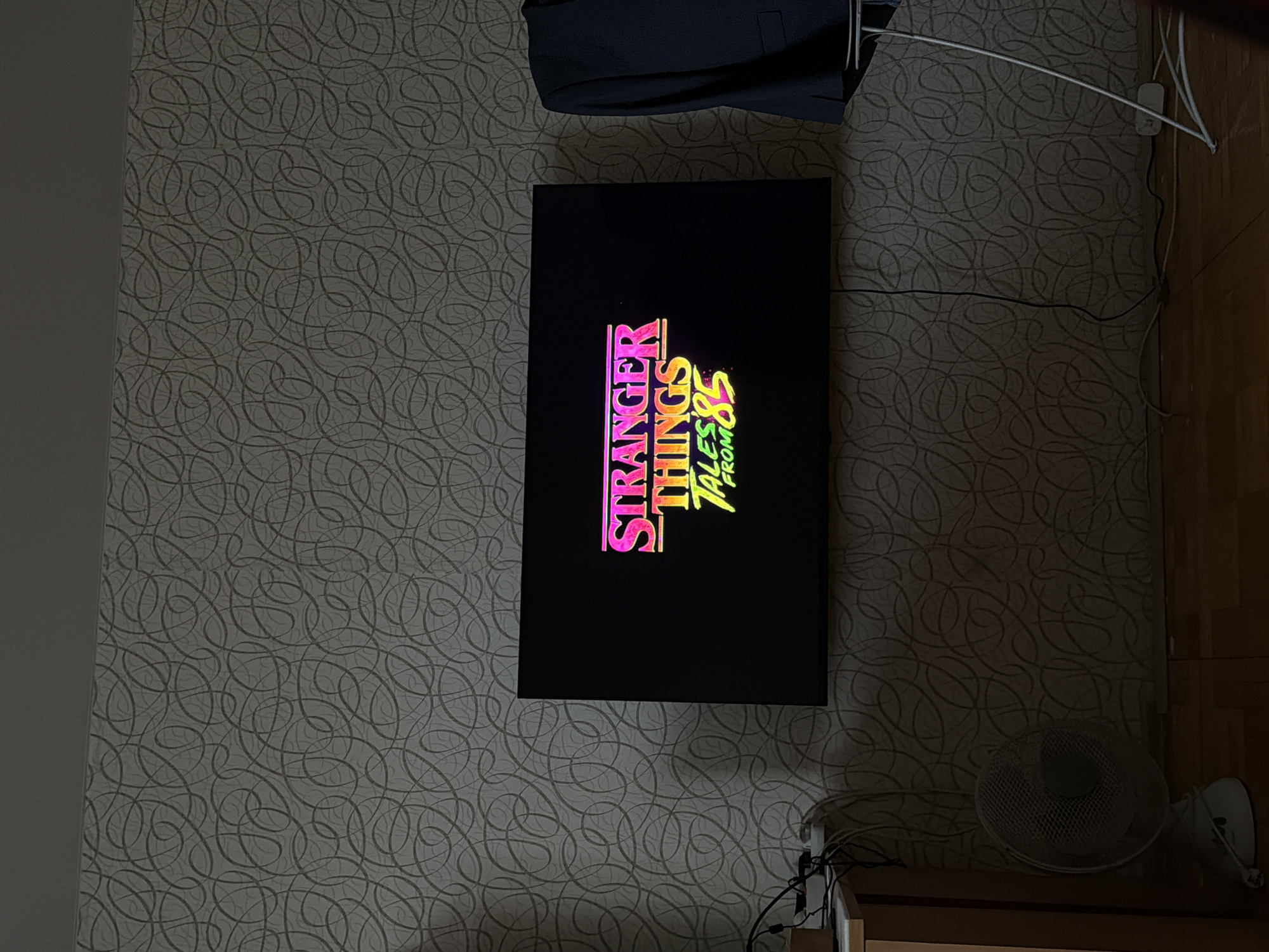 The image displays an interior room, likely a residential space, viewed with a 90-degree counter-clockwise rotation. The central focus is a large, black flat-screen television that is currently powered on and displaying a title screen.
On the television screen, the text "STRANGER THINGS" is prominently shown in a pink and red gradient, using a distinctive 80s-inspired font. Below this, smaller text in yellow and green reads "TALES FROM '85", with the "'85" part styled in a more graphic, almost graffiti-like manner.
The wall behind the television features a light grey wallpaper with a repeating, intricate pattern of swirling dark grey lines. To the left side of the frame, there is a plain, dark grey wall section. Above the television, a dark-colored jacket or piece of clothing is draped.
Below and to the right of the television, a white desk fan with a circular grate is visible, resting on a wooden surface, possibly a shelf or cabinet. Several black and white cables are present, connected to a white power strip or outlet mounted on the wall. These cables snake across the wallpaper.
The room appears dimly lit, suggesting it is either evening, night, or that ambient light sources are limited. No people are visible, and no specific emotions can be discerned. The image provides a glimpse into a personal viewing experience in Perm, Russia.