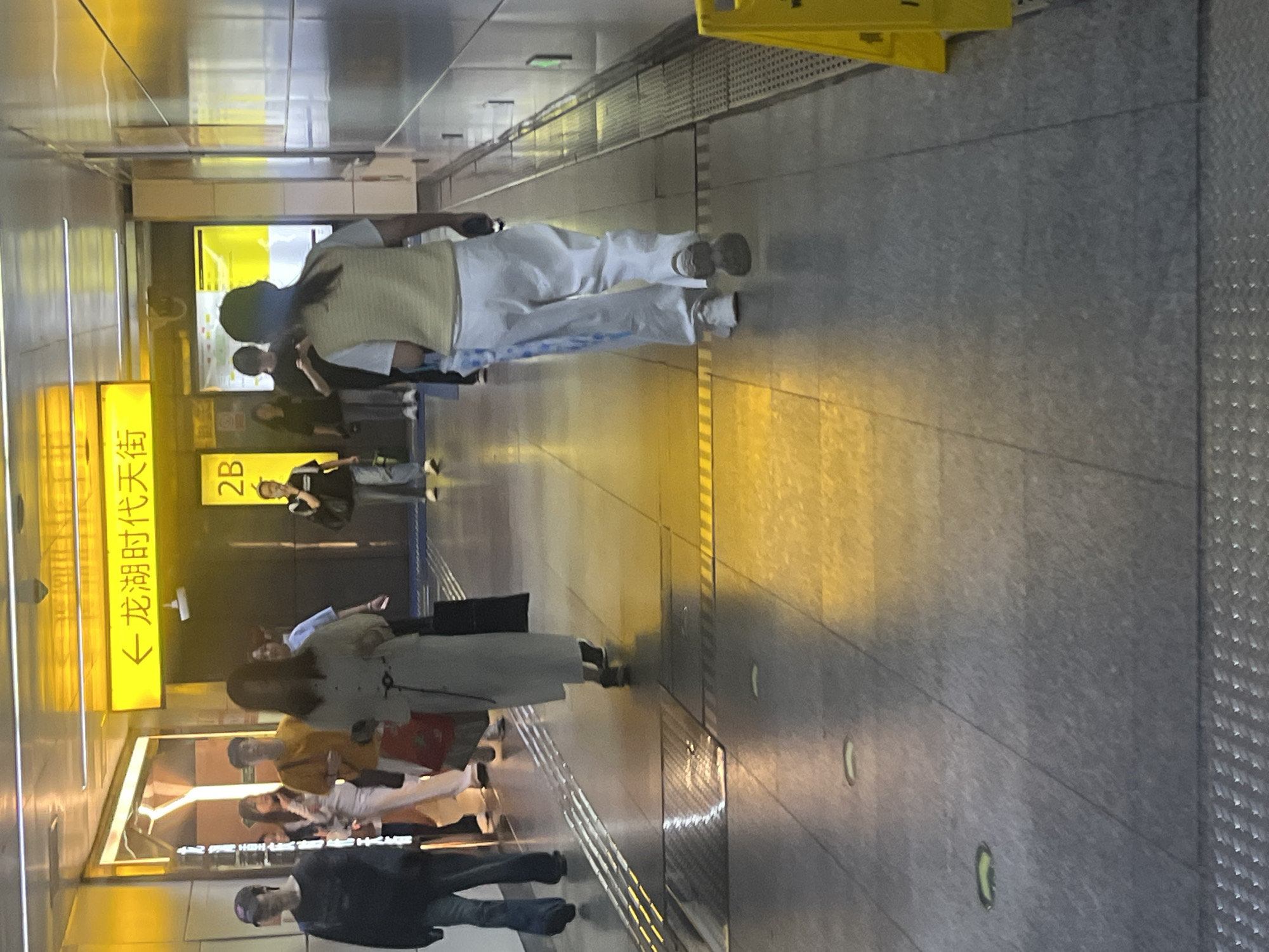 In a brightly lit, modern subway station in Yuzhong District, China, several individuals navigate the concourse. A woman in a light-colored, long coat walks away from the camera, carrying a dark tote bag. To her left, a woman in white pants and a light-colored vest also walks away from the camera. Further ahead and to the left, a man in a dark shirt and cap is visible. To the right of the woman in the white pants, a person in a black t-shirt and light-colored pants walks away from the camera. In the background, a yellow sign with Chinese characters and a directional arrow is illuminated, indicating a destination. The station floor is tiled with a contrasting patterned strip and reflective sections, likely from lighting. A yellow caution sign is visible on the far right.