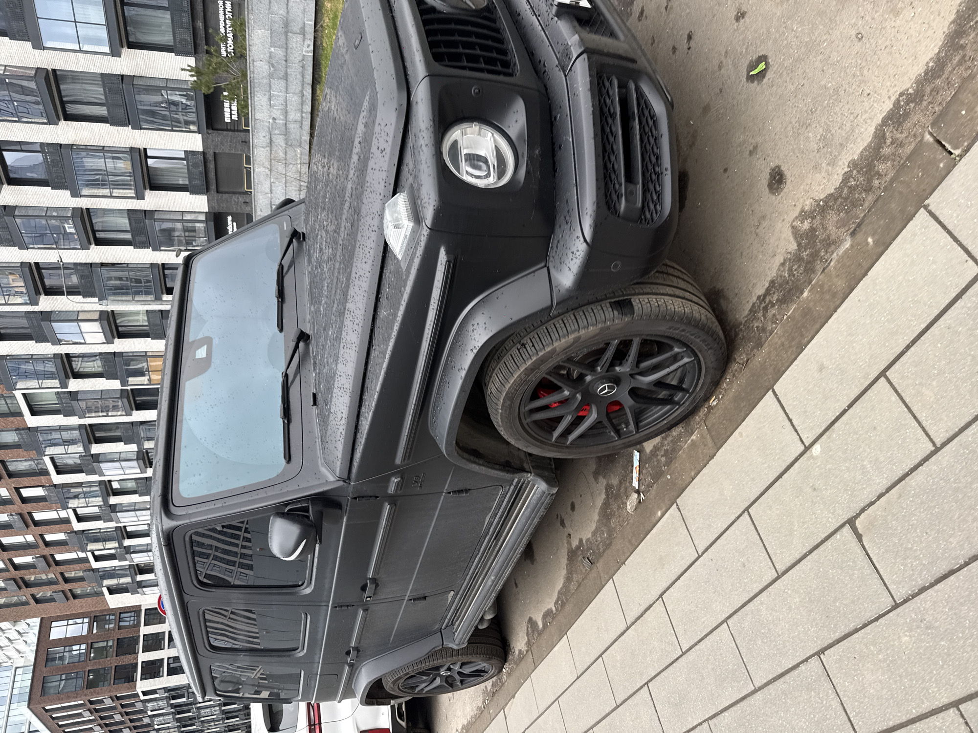 A matte black Mercedes-Benz G-Wagen (G-Class) is parked on a paved surface next to a light-colored tiled sidewalk. The vehicle's exterior, including the hood and roof, is covered in water droplets, suggesting recent rain or damp weather. It features black multi-spoke wheels with prominent red brake calipers, and robust side steps are visible along its lower side profile. The front grille, round headlights, and square turn signals are characteristic of the G-Wagen model.
In the background, a modern multi-story building with a facade alternating between light-colored brick and darker sections, featuring numerous windows and balconies, stands tall. Some green foliage is visible near the base of the building. The sky appears overcast, indicating daytime.
Cyrillic text is visible on the building's signs. One sign clearly reads "ЦЕНТР СОВРЕМЕННОЙ СТОМАТОЛОГИИ" (Center of Modern Dentistry), while another below it states "ПИВНАЯ ЛАВКА" (Beer Shop). Partially visible words like "Vandome" and "Отель" (Hotel) can also be seen. The setting is an urban or suburban area, consistent with Khimki, Russia. No people are present in the image, and the car is stationary.