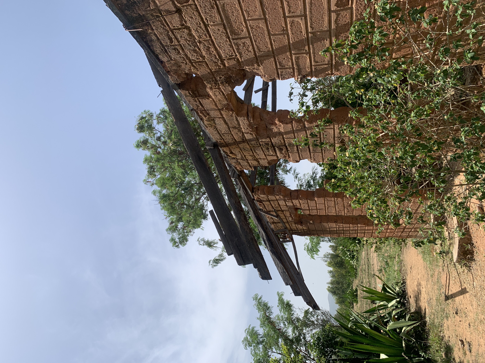 The image shows a dilapidated brick structure in an outdoor setting, possibly located in Gwafan, Nigeria. The structure's roof is partially collapsed, with exposed wooden beams. Vegetation, including trees and bushes, surrounds the building, and there's open, grassy land in the background. The sky is visible through the roof opening.