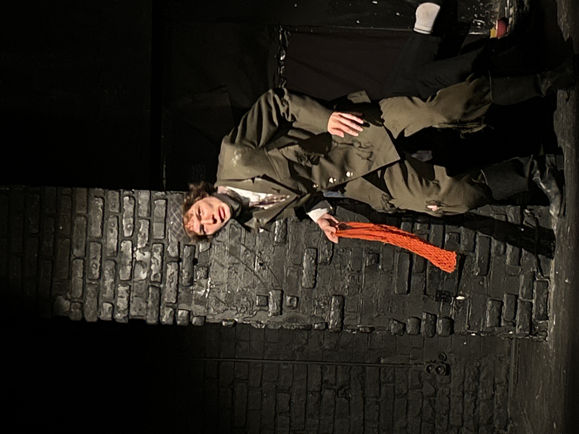 A performer lies prone atop a dark, faux-brick structure. The figure is attired in a greenish-brown jacket and matching trousers. A patterned cap rests on their head, and they are holding an orange scarf. The setting appears to be a theatrical stage, with black backdrops framing the scene. The location seems indeterminate, as the artificial brickwork does not suggest a specific city or place.