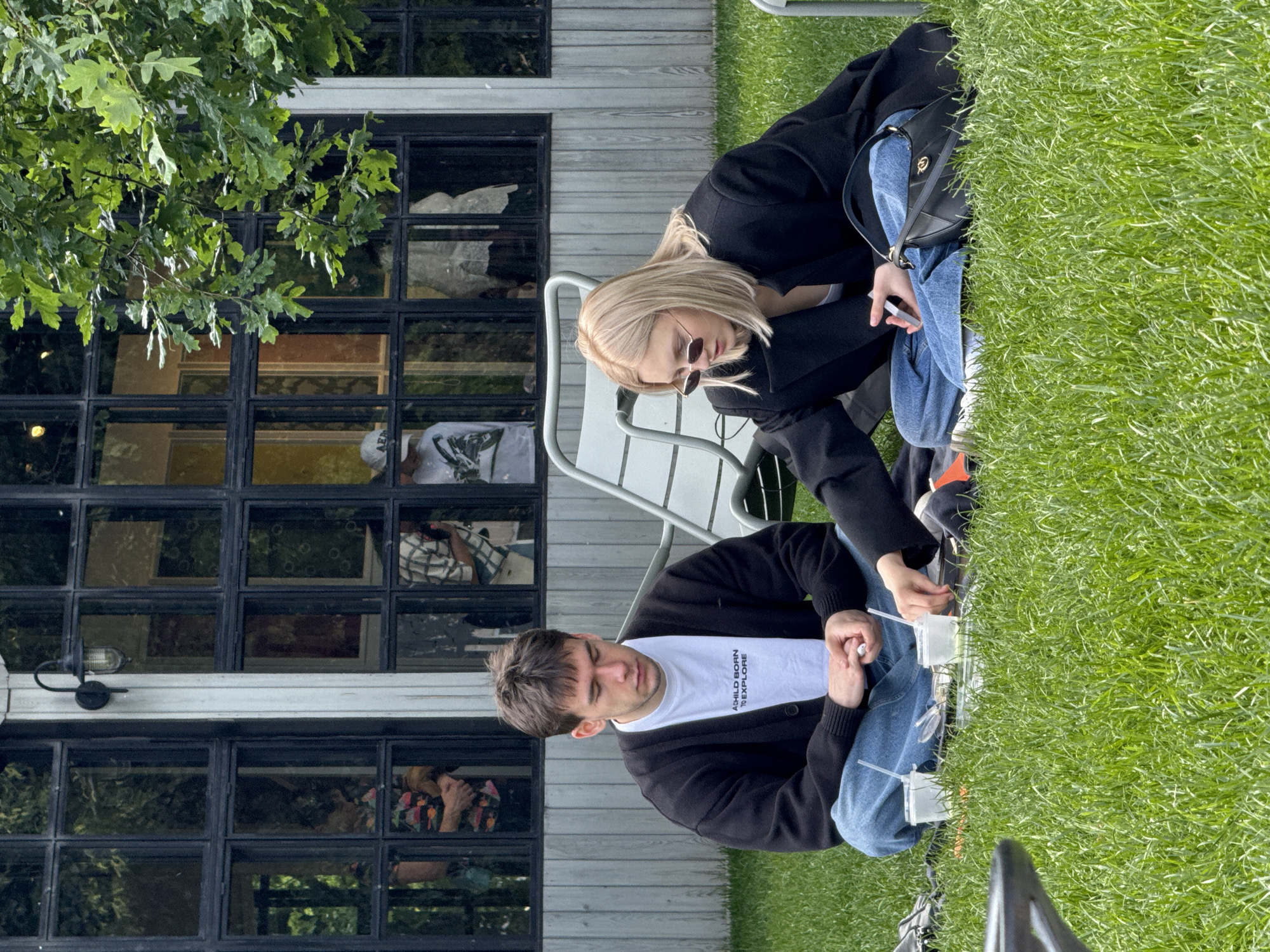 The image depicts a young man and a young woman sitting on the grass in front of a building with large windows, suggesting an outdoor cafe or restaurant in Saint Petersburg, Russia. The man is wearing a black cardigan over a white t-shirt and blue jeans. He is sitting cross-legged with his hands clasped in his lap, eyes closed, appearing contemplative. The woman, with blonde hair and sunglasses, is wearing a black blazer over a white top and blue jeans. She is looking down at her phone. Between them, on a picnic blanket, are two clear plastic cups with straws, a small tray with food, and other items. In the background, through the windows of the building, blurred figures and reflections are visible. A large tree with green leaves partially obscures the upper right portion of the building. The overall setting suggests a casual outdoor gathering in a green space within an urban environment.