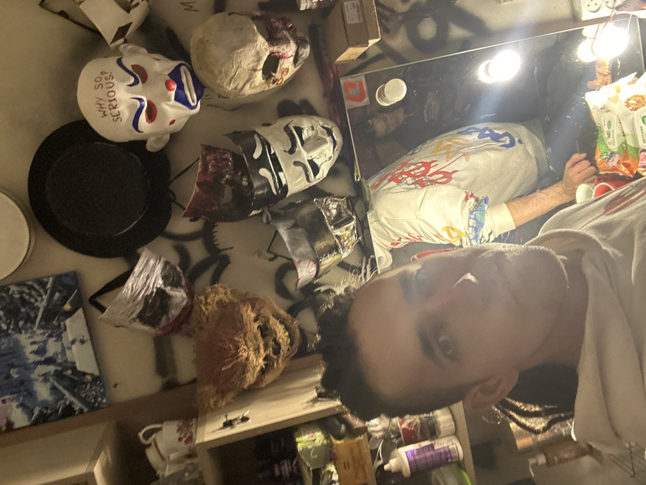 The image captures an indoor scene in Saratov, Russia, featuring a male subject taking a selfie in a cluttered room. The primary subject, positioned in the lower-left foreground, is a male with dark hair styled with shaved sides and a textured, possibly dreadlocked, top and back. He wears a light-colored hooded sweatshirt and looks directly into the camera.
The wall behind the subject is light-colored with extensive dark graffiti. Numerous theatrical or costume masks are affixed to the wall. These include: a white mask with red "WHY SO SERIOUS?" text, depicting a clown-like face with red eyebrows, red eye outlines, a blue nose, and a blue downturned mouth; a pale, distressed mask with deep eye sockets and reddish-brown stains, resembling a skull or zombie; a large, light-brown, furry monster mask with exposed teeth; and several other masks, including one dark red, one metallic silver, and a white mask featuring black comedy/tragedy-style eyes and mouth. A black fedora-style hat hangs near the top center of the wall, and a framed, blue-toned artwork depicting a cityscape is visible on the upper-left.
To the left, a light-colored wooden shelving unit displays several white and red ceramic mugs on its top shelf, with various bottles and containers, including one with a prominent purple label, on lower shelves.
On the right side of the image, a large rectangular mirror is partially visible. Its reflection shows a second individual, a person with dark hair wearing a dark top, sitting or standing further back and looking at a mobile phone. Also visible in the reflection on a surface in front of the mirror are several red plastic cups, a clear glass, and multiple plastic bags containing snacks or goods, some with Cyrillic labeling (e.g., "ДЕТСКИЕ"). Two circular lights are attached to the right frame of the mirror, illuminating the reflected area.