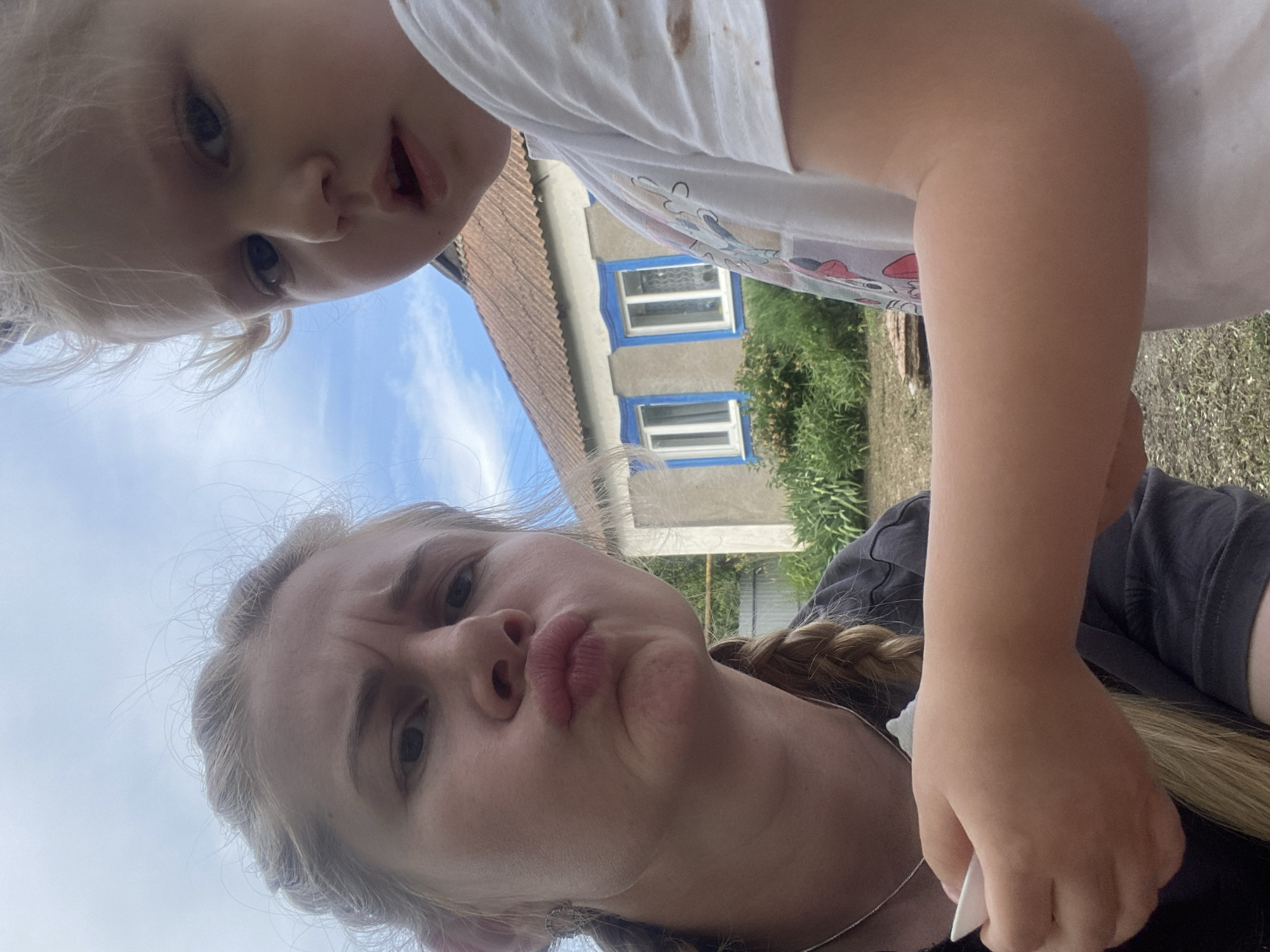 The image is a selfie taken outdoors, with two subjects visible: a young child and a woman. The child is in the foreground, looking directly at the camera with wide eyes and an open mouth. The woman is positioned below the child, also looking at the camera with a playful, puckered expression. Her hair is styled in a braid.
The background features a building with a tiled roof and windows with blue trim. Green foliage and grass are also visible, suggesting a rural or suburban setting. The sky above is blue with some clouds, indicating daylight.
The woman appears to be holding a white object in her hand, possibly a phone or a similar device, as she is taking the photo. The overall mood of the image is lighthearted and casual, likely a candid moment captured between the two individuals. No text is visible in the image.