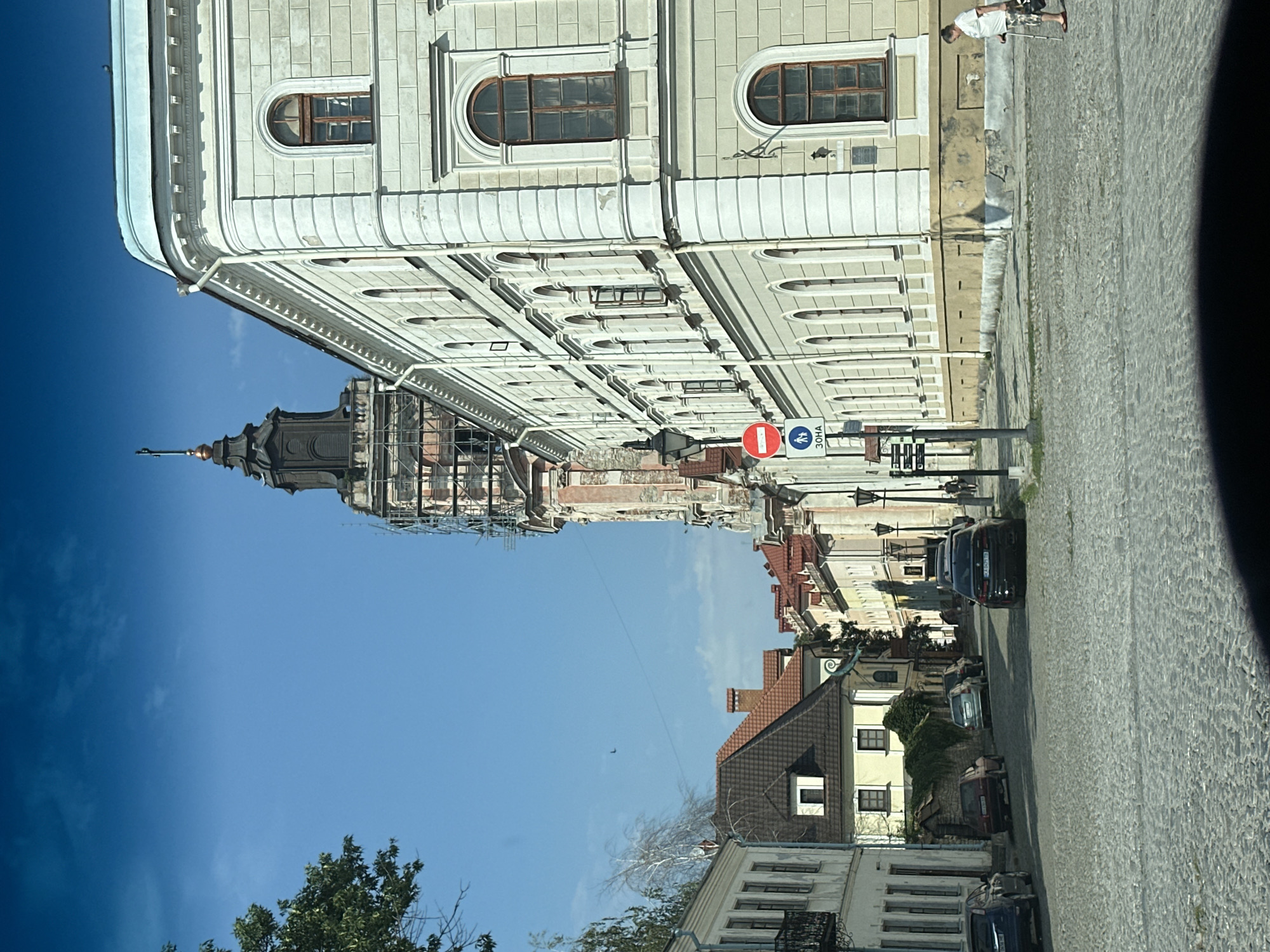 The image depicts a street scene in Kamianets-Podilskyi, Ukraine. The architecture features historic buildings with neoclassical elements, characterized by their light-colored facades, arched windows, and detailed cornices. A prominent tower, partially obscured by scaffolding, dominates the background, suggesting ongoing construction or renovation. Cobblestone paving covers the street, and several cars are parked or driving. Pedestrians are visible, including a man with a cane near the right side of the frame. Traffic signs, including a red circular prohibition sign and a green sign with pedestrian figures and Cyrillic text, are present. The sky is clear and blue, indicating fair weather. The overall scene captures a blend of historical preservation and daily urban life in the city.