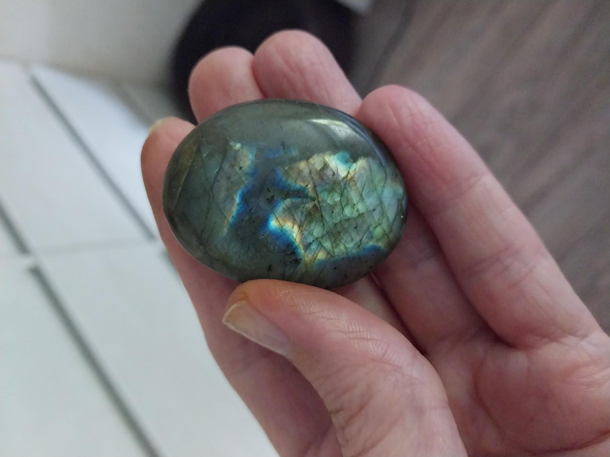 An oval-shaped, polished stone, possibly labradorite due to its iridescent blue and green flashes, is held in a person's hand. The stone has a dark grey to green base with distinct crack-like inclusions. The hand appears to be pale-skinned, with visible creases and a fingernail on the index finger. The background is blurred, showing white horizontal lines which could be part of furniture or a wall, and a wooden floor. There is no indication of the location being in Dnipro, Ukraine.