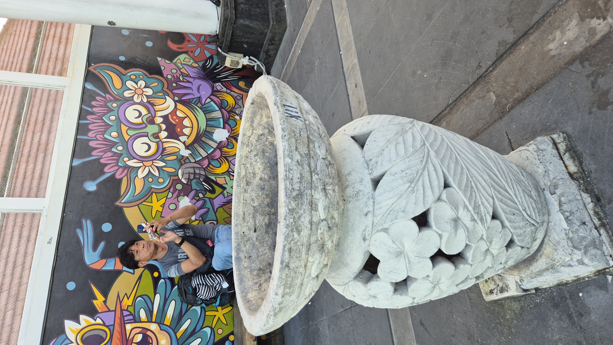 A medium shot, taken outdoors, depicts a man seated beside a large, weathered stone urn in Kuta, Indonesia. The man is positioned against a vibrantly colored mural, with blues, greens, yellows, pinks, and purples forming abstract and floral designs. He holds a small item in his hands, possibly a camera or phone, which he appears to be using. The stone urn, composed of two pieces, is carved with decorative leaves and flowers. The base rests on a square stone platform. The ground is paved with gray concrete, which is cracked in places. A section of a building with a window is visible in the background, and black trim runs across the upper portion of the mural.