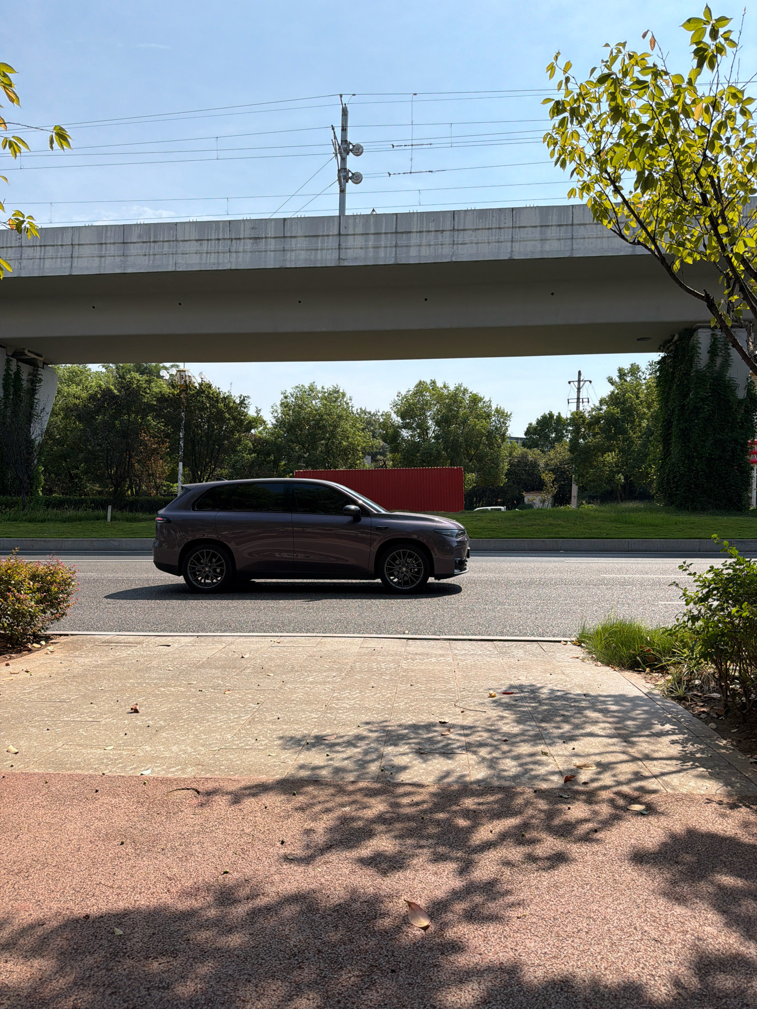 A gray SUV is driving on a road under an elevated highway or bridge in Jiangjin, China. The highway is supported by concrete pillars and has overhead power lines. Trees and green spaces are visible on either side of the road, with a red fence in the background. The foreground shows a paved pedestrian walkway with dappled sunlight and shadows from overhead trees. The sky is clear and blue.
